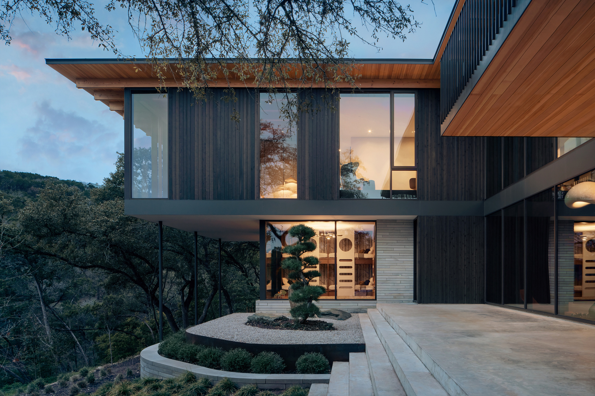 Dark timber-clad upper floor over a lit ground level, with a sculpted pine in a gravel bed at dusk