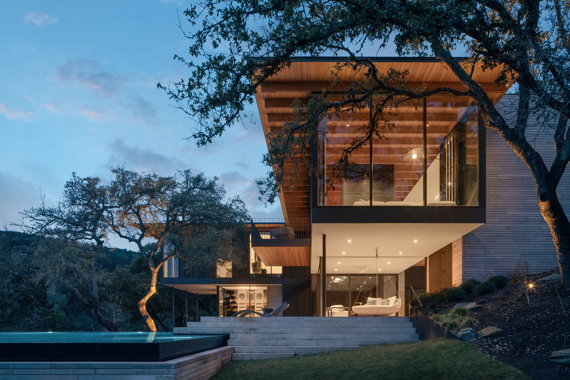 Cantilevered glass upper volume with timber soffit above a stone stair and infinity pool, framed by live oaks at dusk