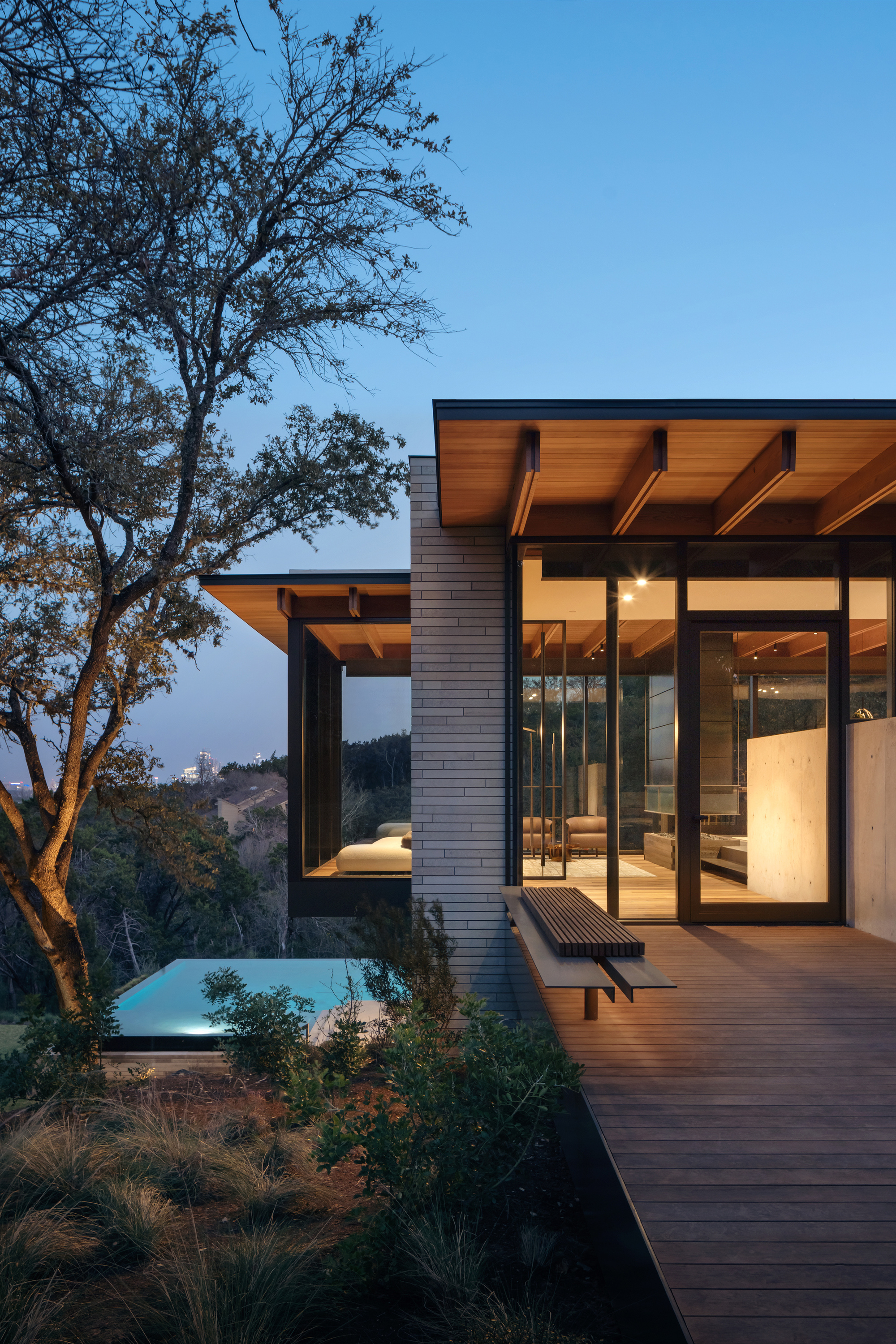 Timber-roofed pavilion with floor-to-ceiling glazing and a slatted bench on a wooden deck, a lit pool below at twilight