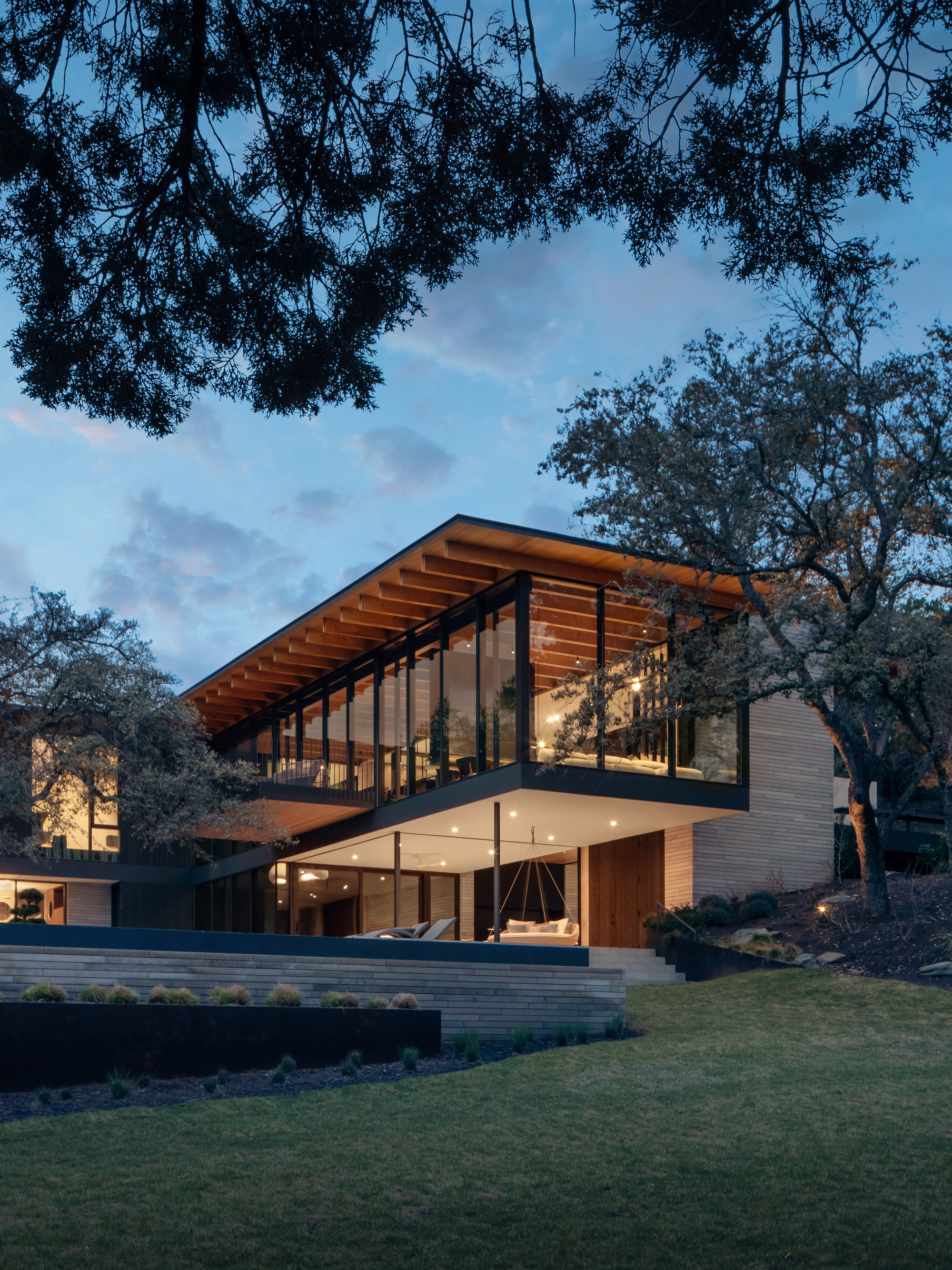 Two-storey glass and timber house with a deep roof overhang, seen across a lawn beneath oak branches at dusk