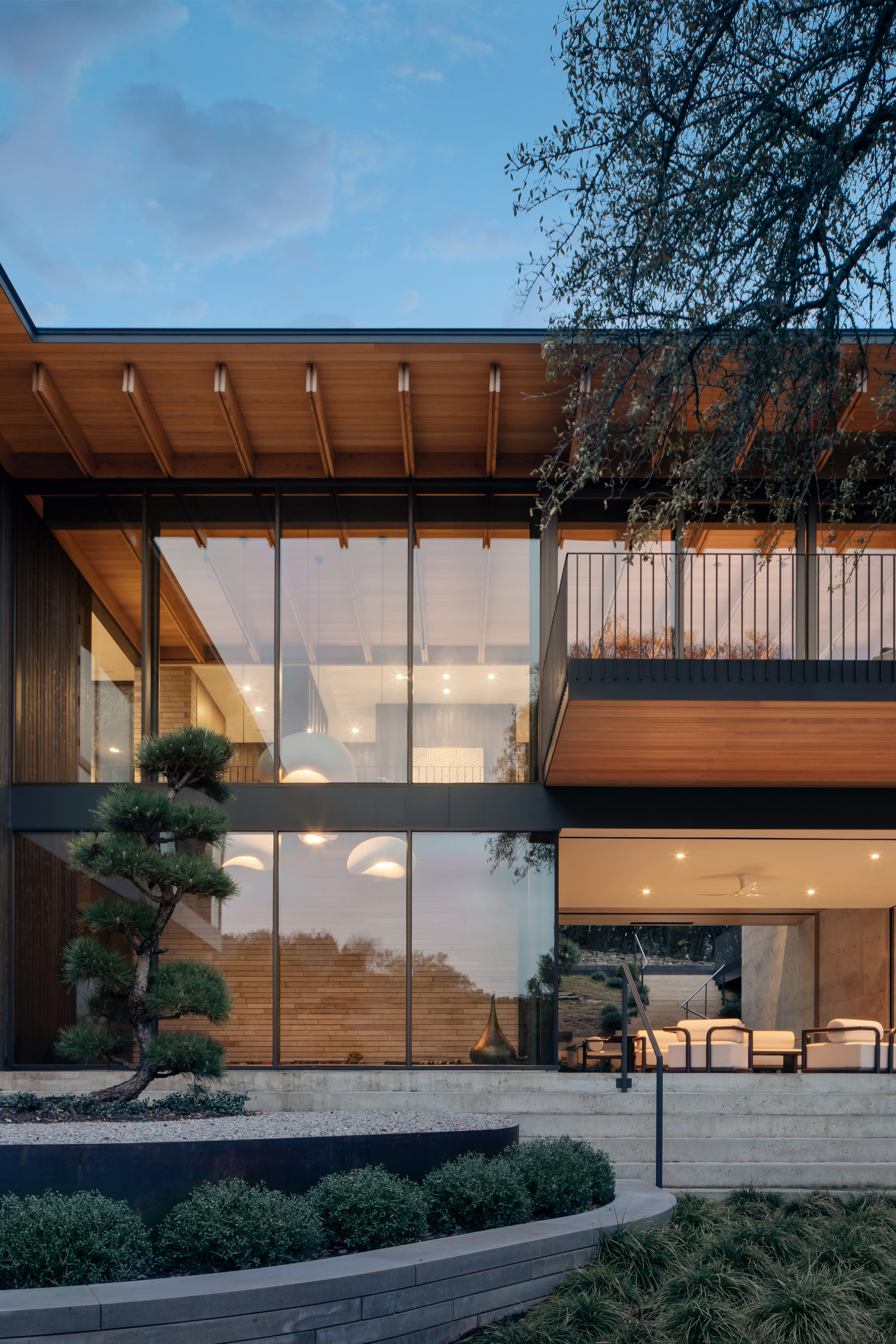 Glazed facade with exposed rafters and a balcony above a terrace, a sculpted pine in the foreground at dusk
