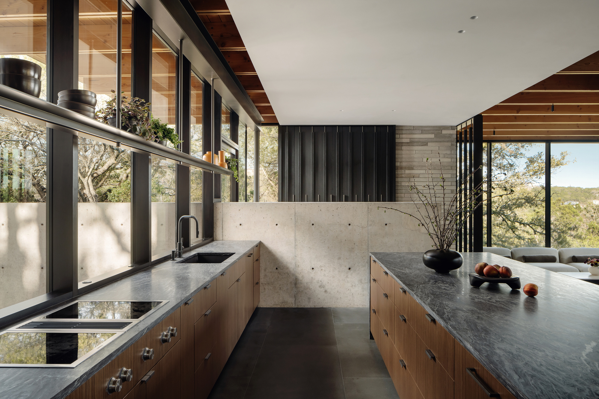Kitchen with walnut cabinets, dark stone counters and a board-formed concrete wall beside a continuous window band looking onto trees