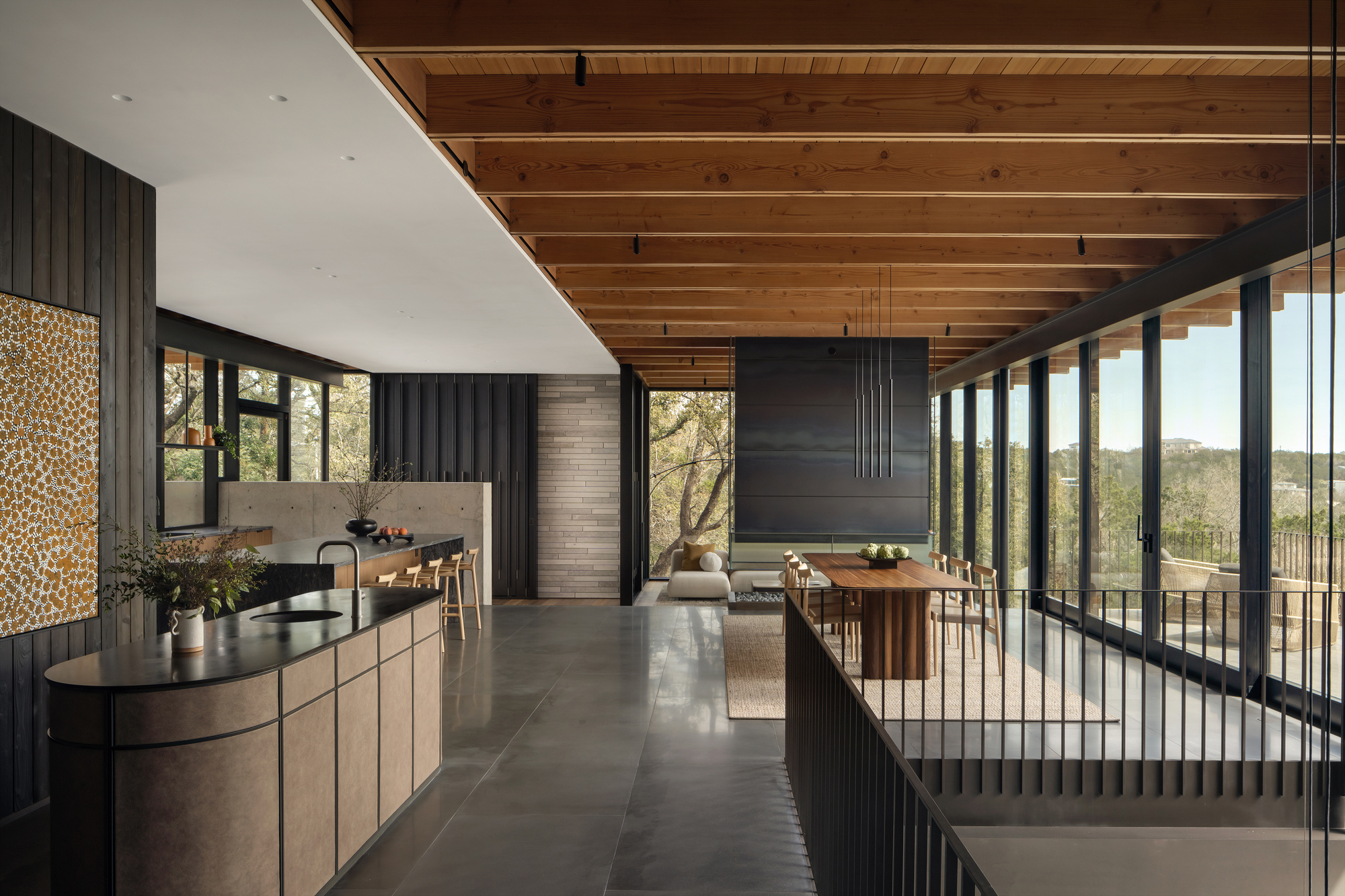 Open living space with a curved kitchen island, exposed timber beams and a dining table beside a glass wall facing the treetops