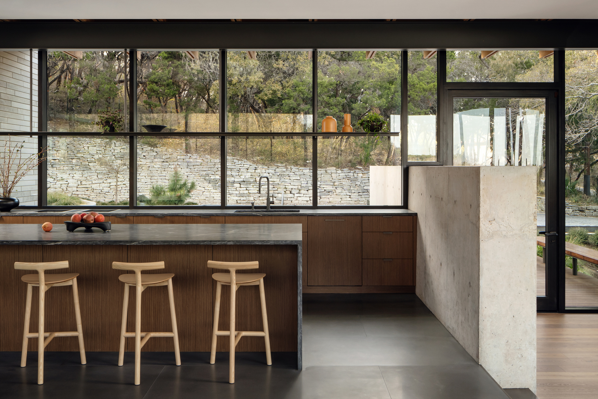 Kitchen island with wood-fronted base and three oak stools facing a glass wall onto a dry-stacked stone retaining wall