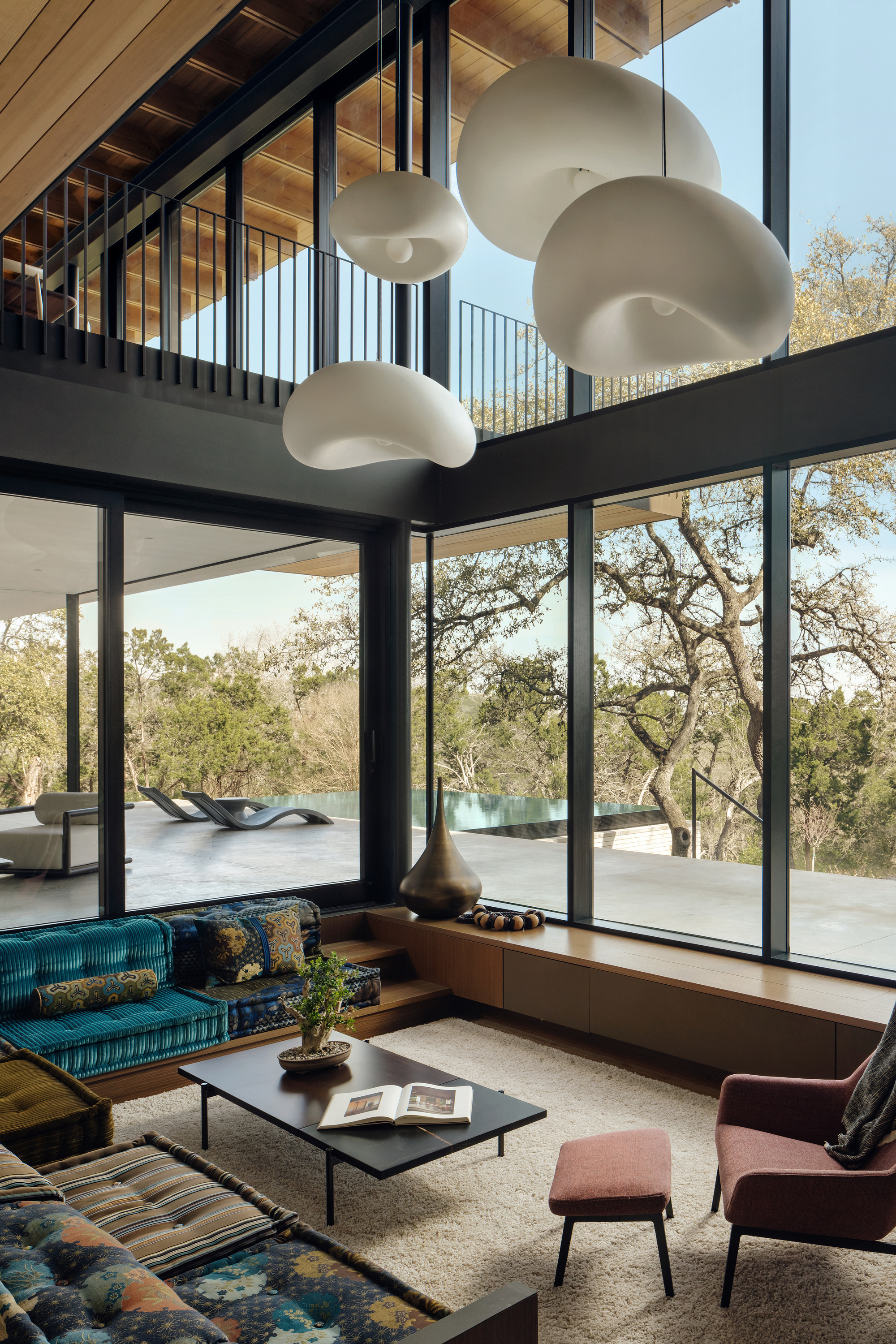 Double-height living room with cloud-shaped pendant lights, teal banquette and floor-to-ceiling glazing opening to oak trees and a terrace