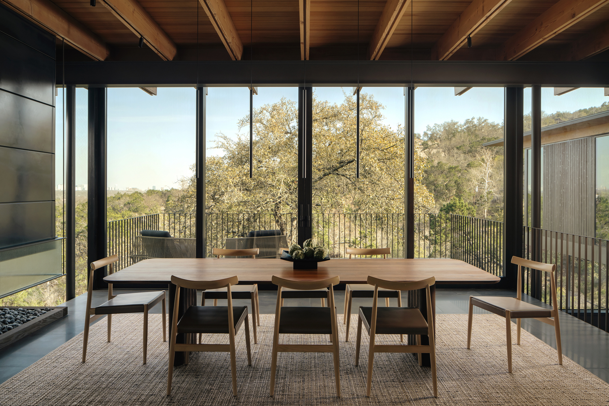 Dining table with oak chairs on a jute rug, beneath timber beams and glass walls framing a live oak canopy