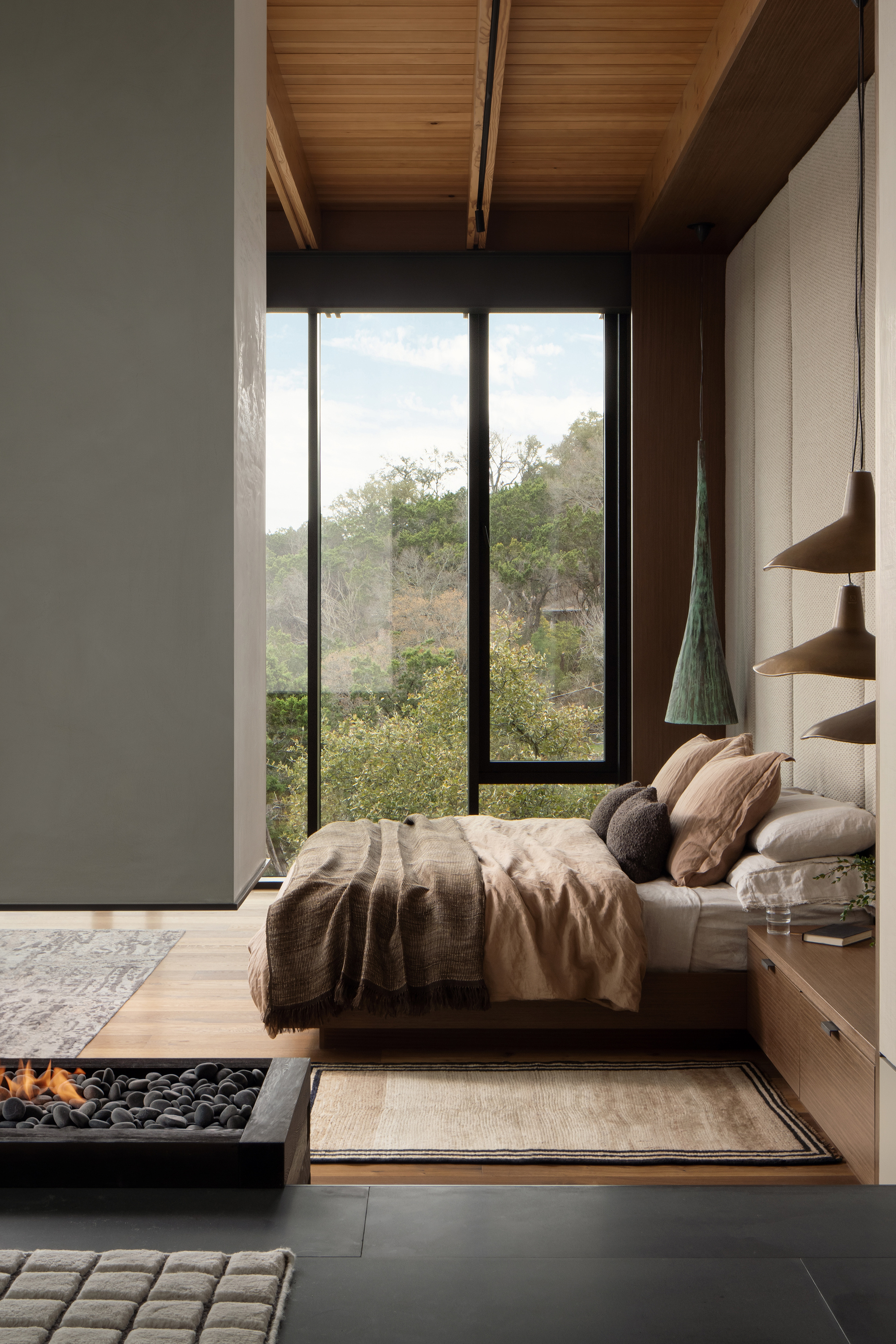 Bedroom with a low platform bed beside a pebble-filled fire trough, facing a tall window onto green treetops