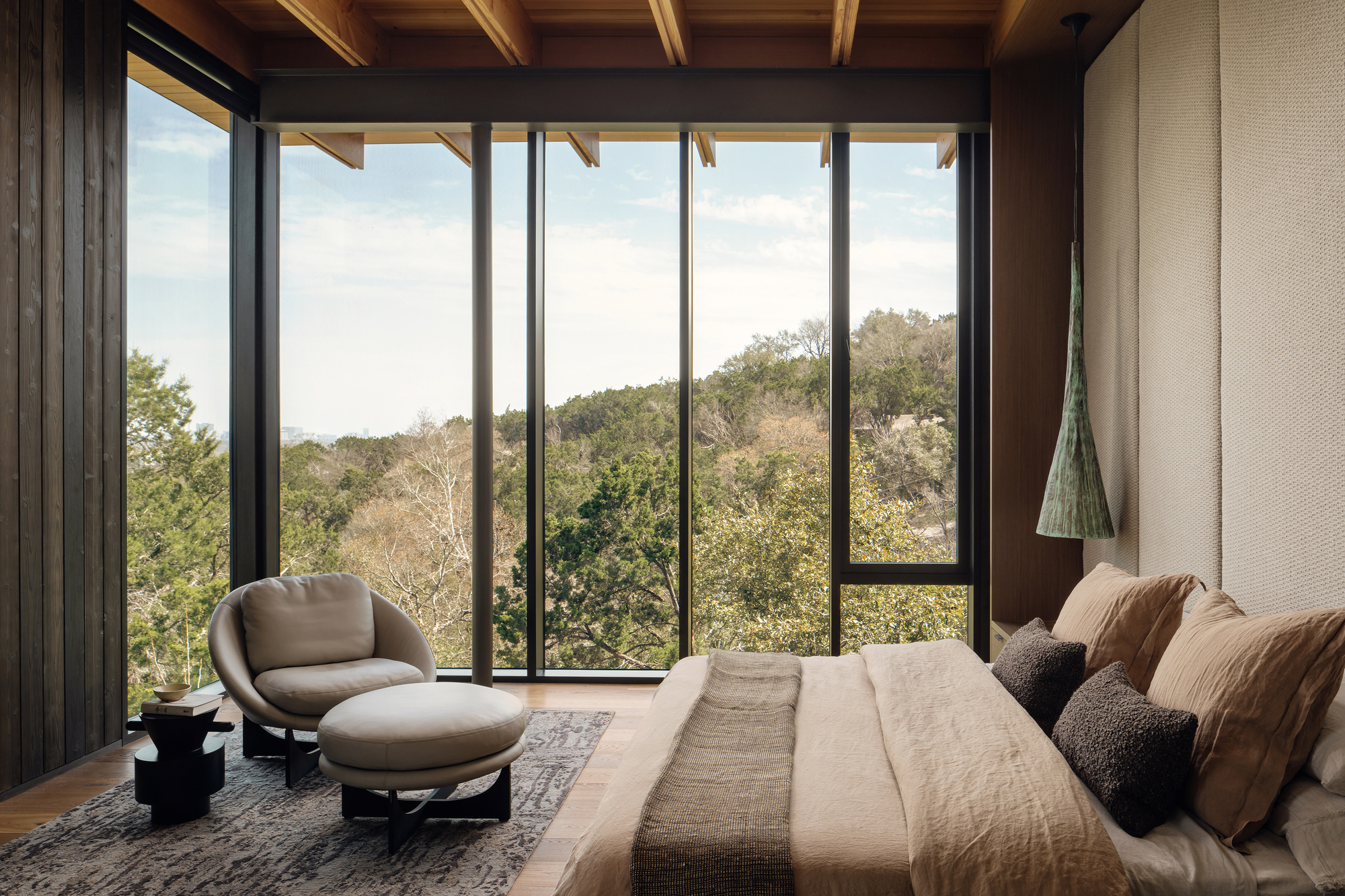Corner bedroom with a round lounge chair and ottoman, wall of glazing and upholstered headboard panels facing a hillside of trees