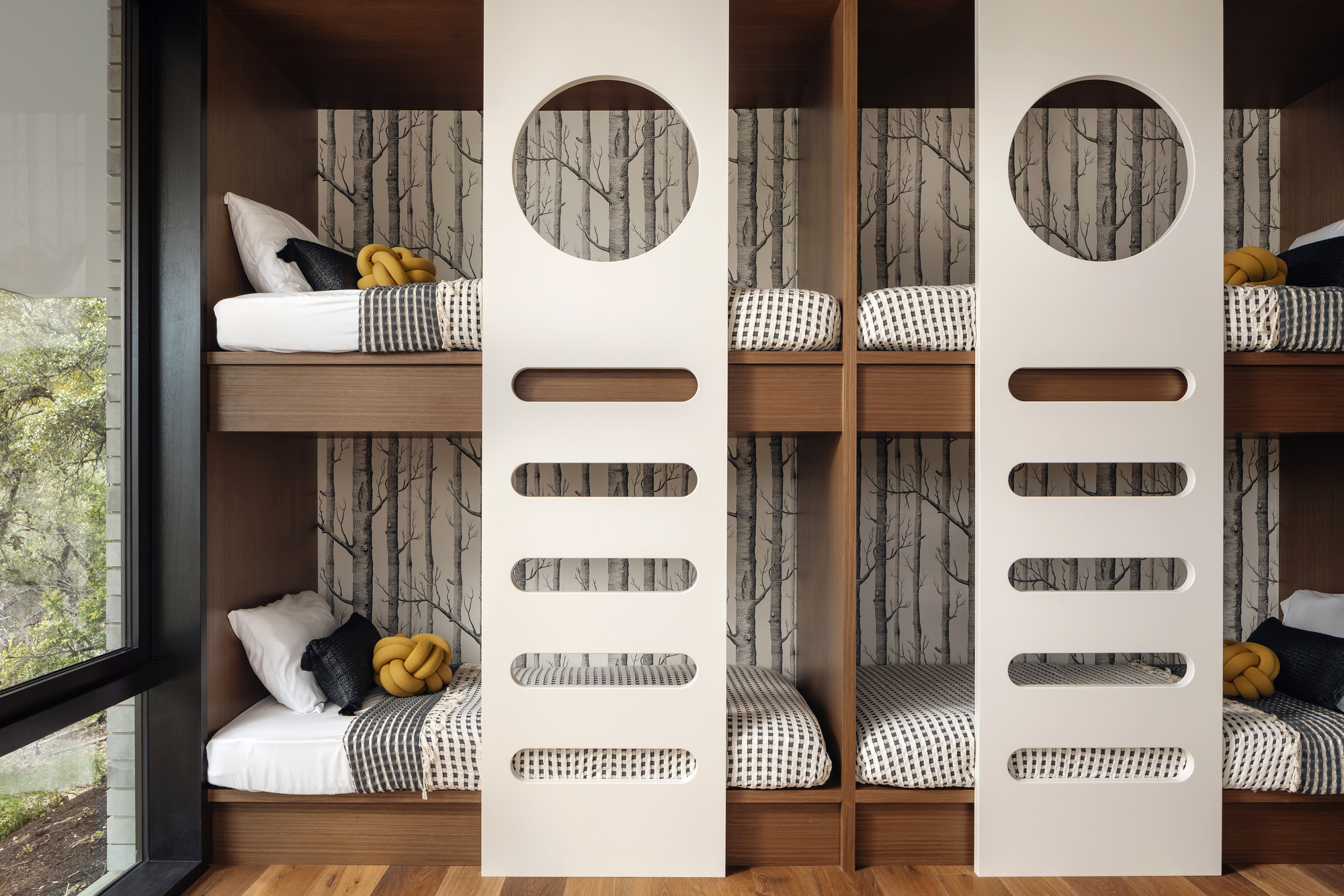 Built-in bunk beds with white ladder panels, circular cutouts and birch-tree wallpaper beside a black-framed window