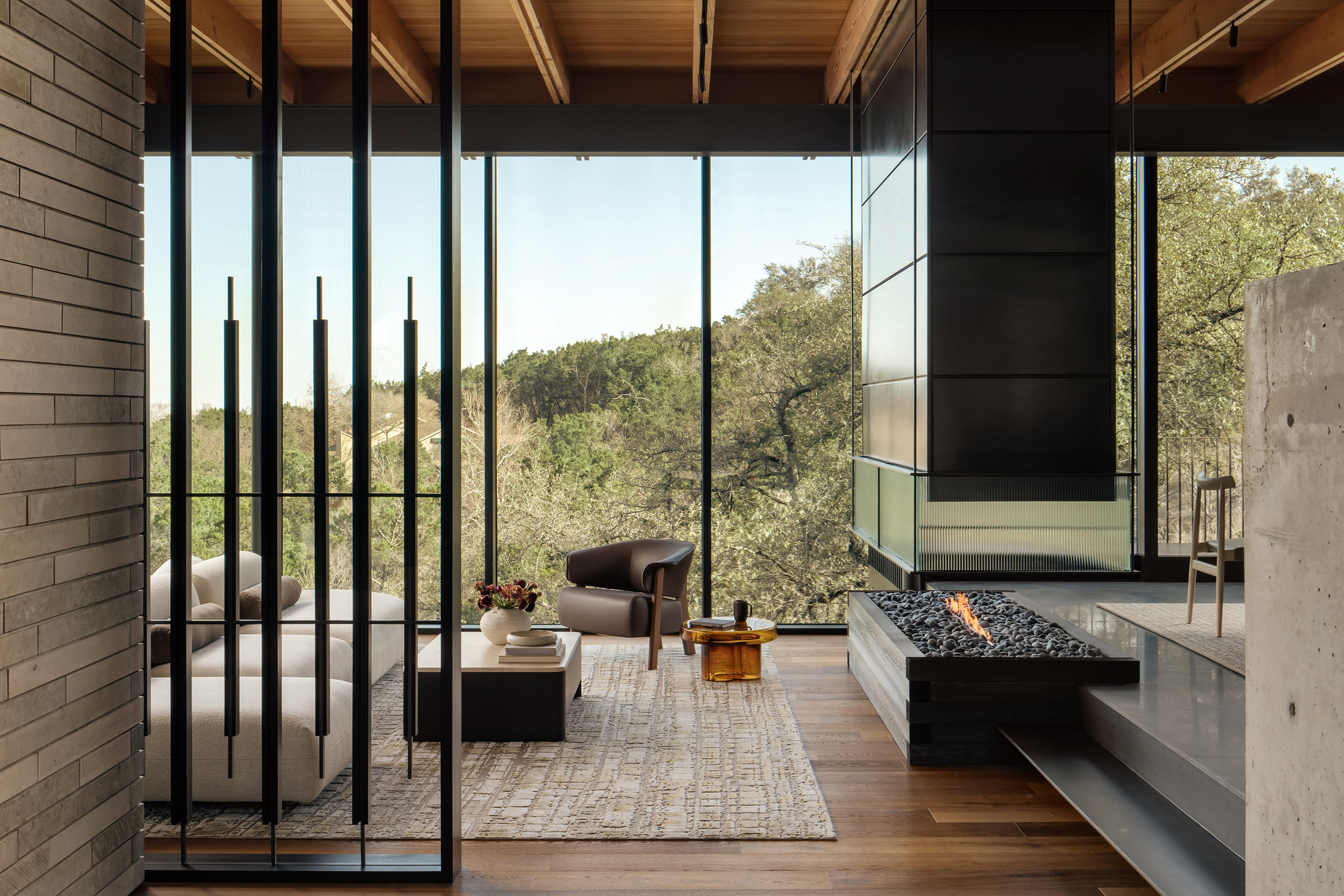 Living area with a linear gas fire pit, black steel screen and floor-to-ceiling glazing framing a wooded hillside in clear daylight