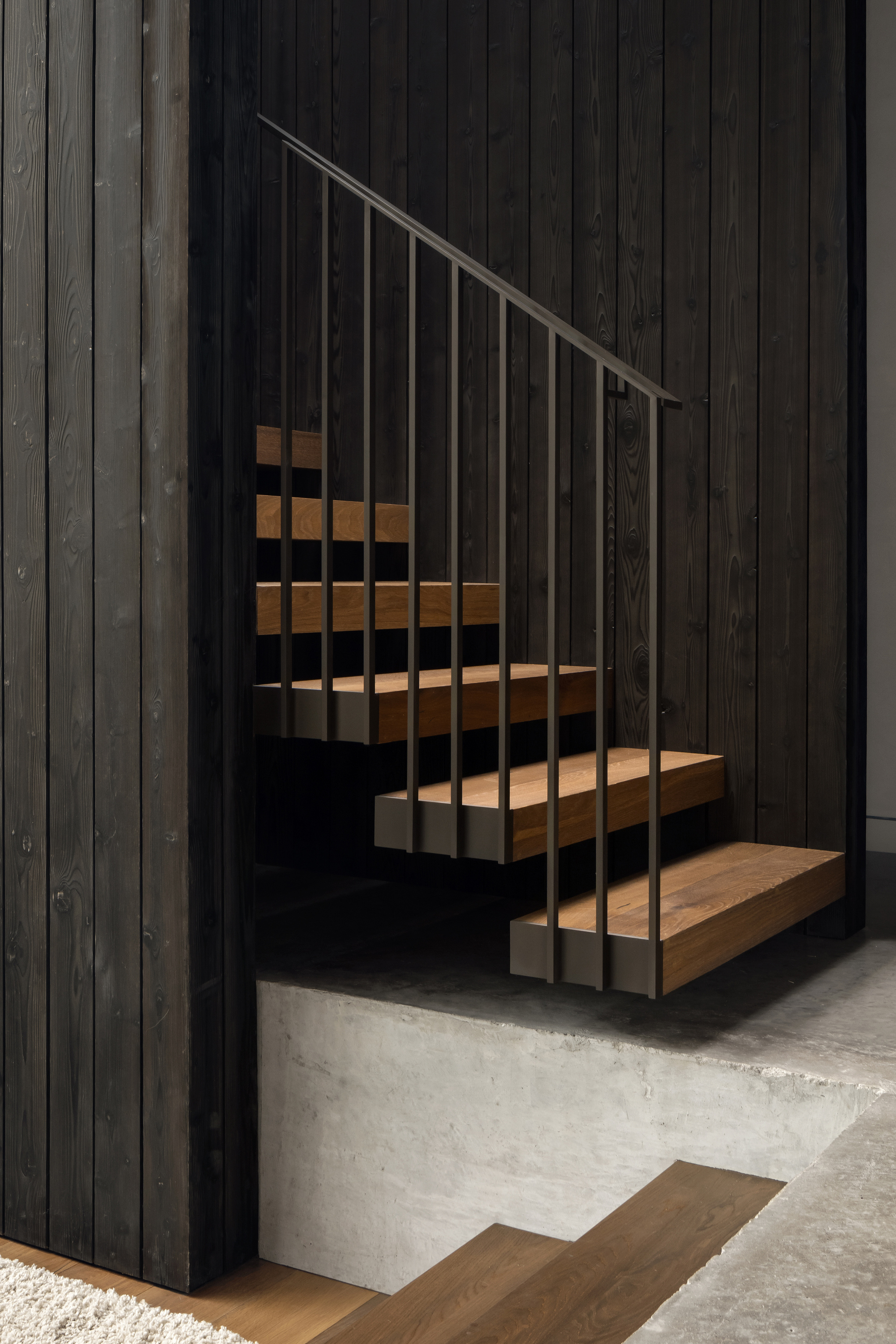 Floating timber stair treads with a slim steel balustrade against charred black vertical wood cladding and a raw concrete plinth