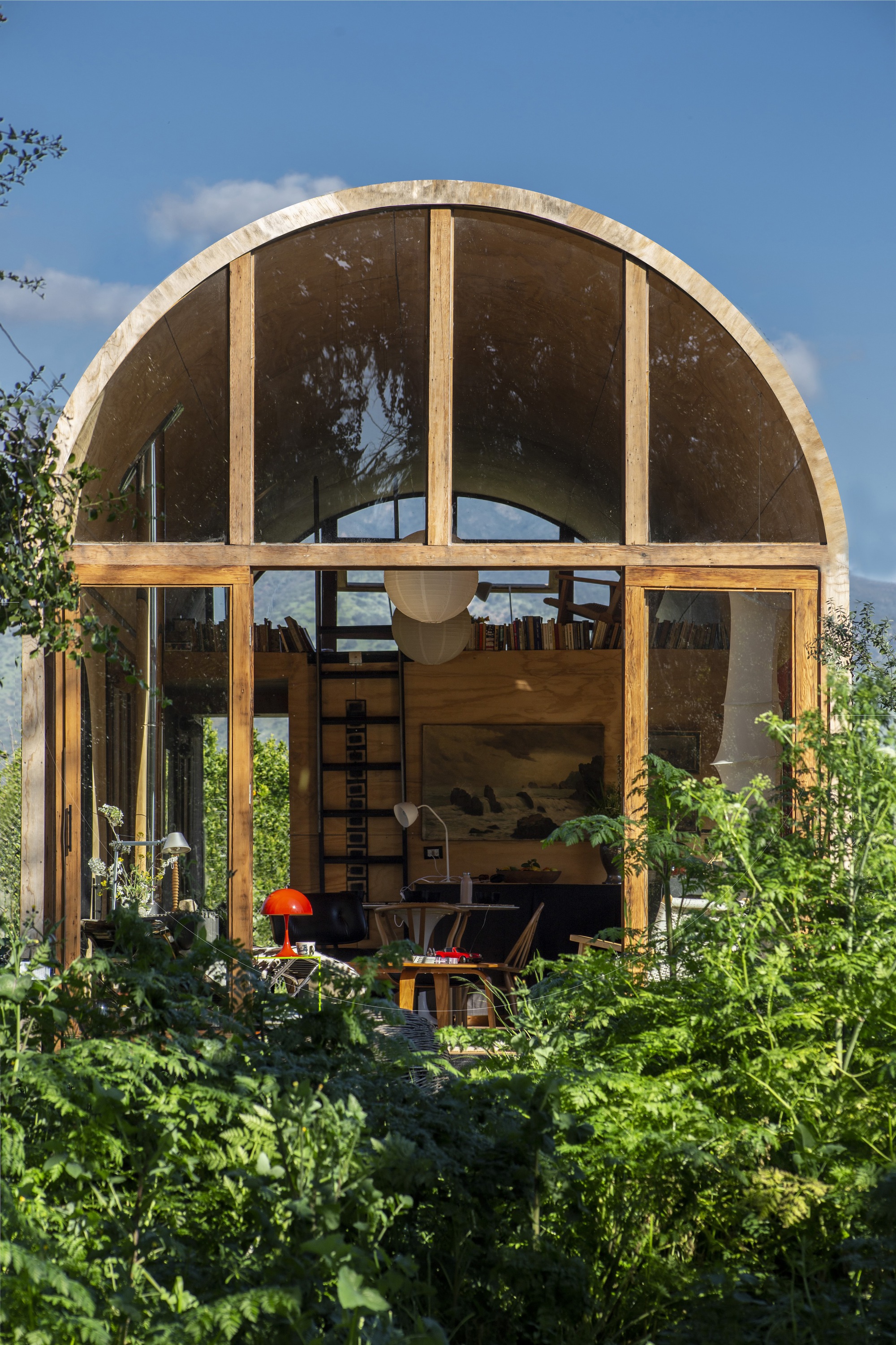 Arched timber-framed glass gable end seen through ferns, revealing a loft ladder, bookshelves and an orange lamp under blue sky