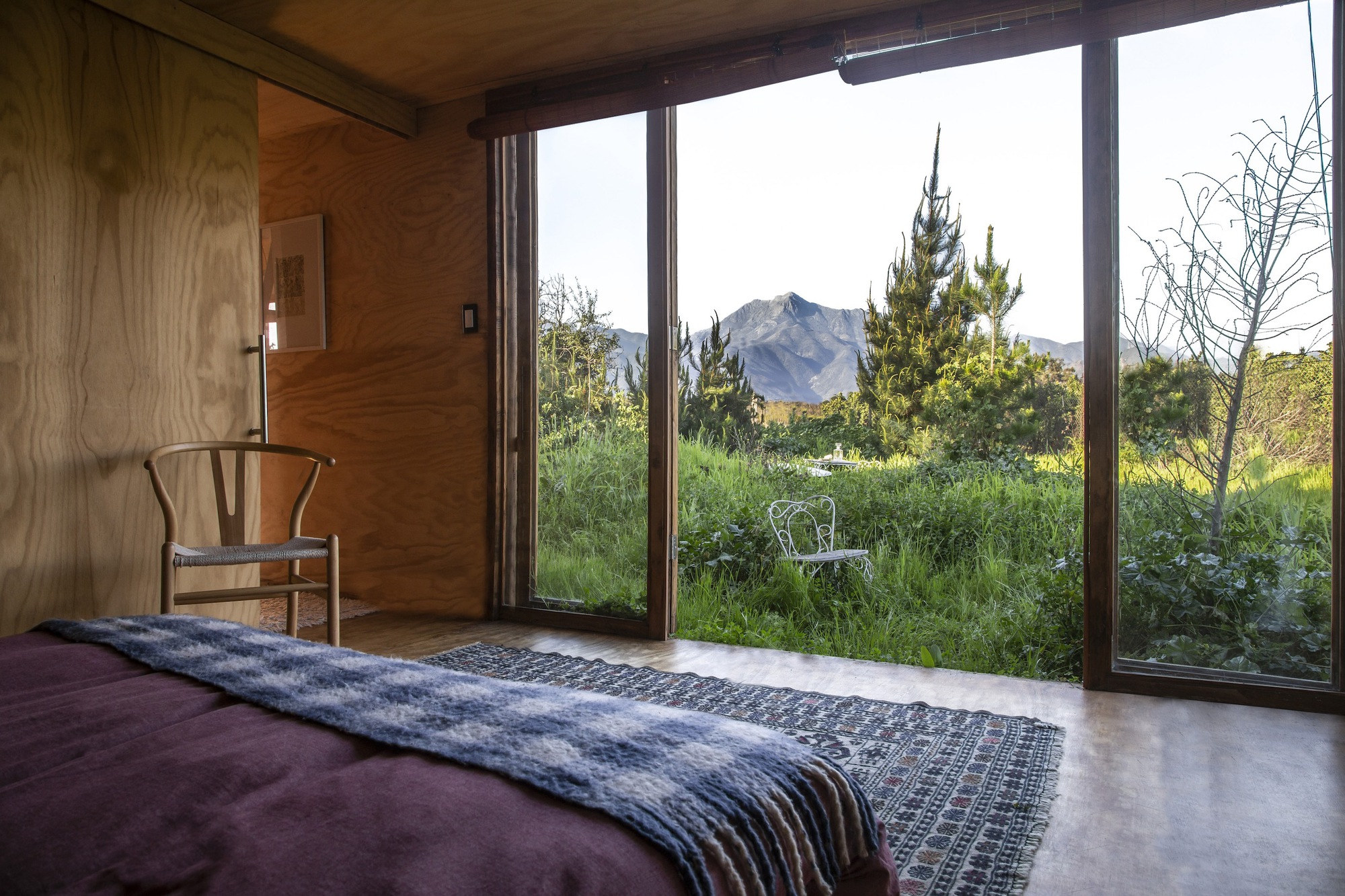 Bedroom with a floor-to-ceiling glass wall opening onto a meadow, pine trees and a distant mountain peak in clear daylight