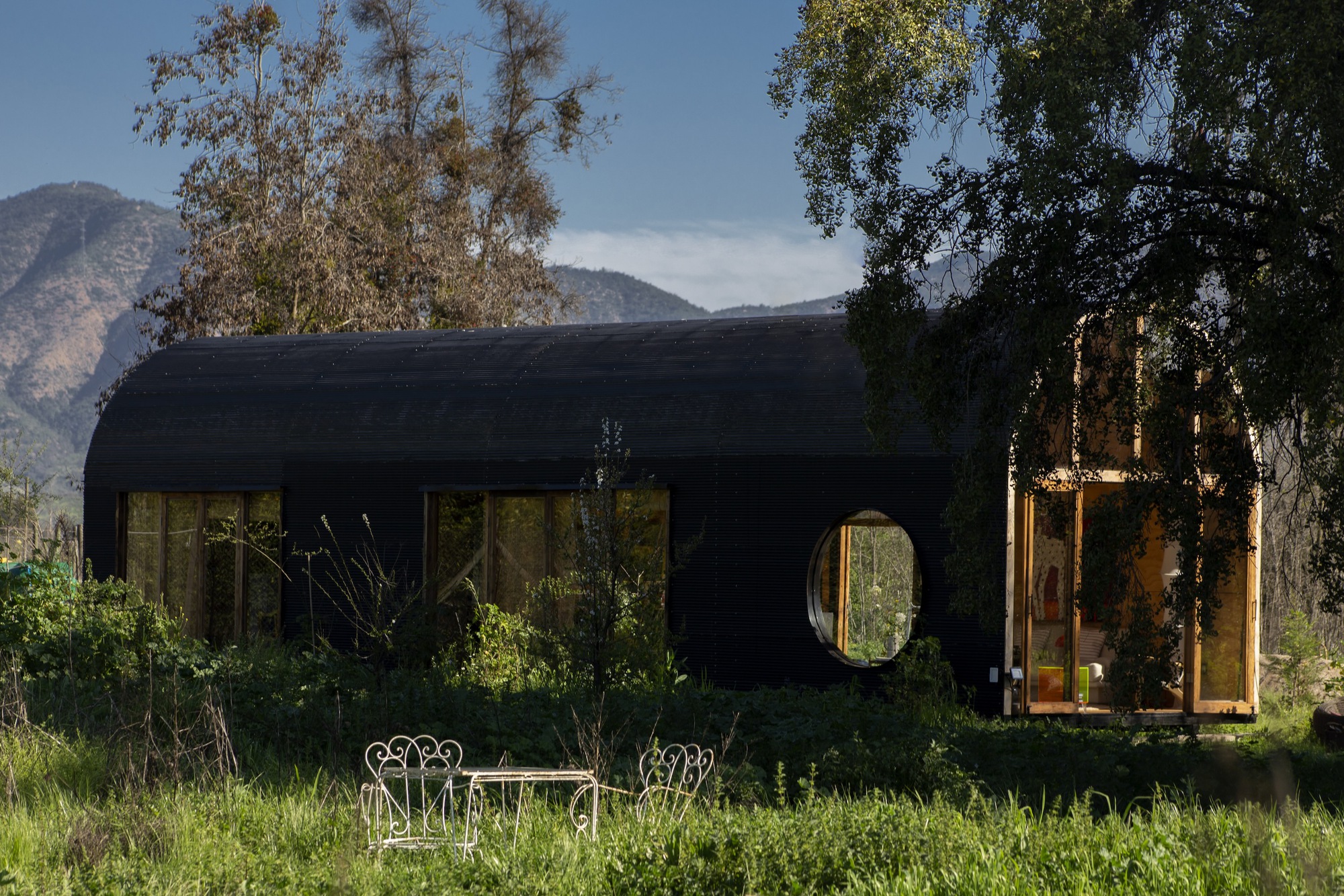 Dark barrel-roofed cabin with a round window and glazed timber end, seen across a meadow with white iron chairs under mountain sky