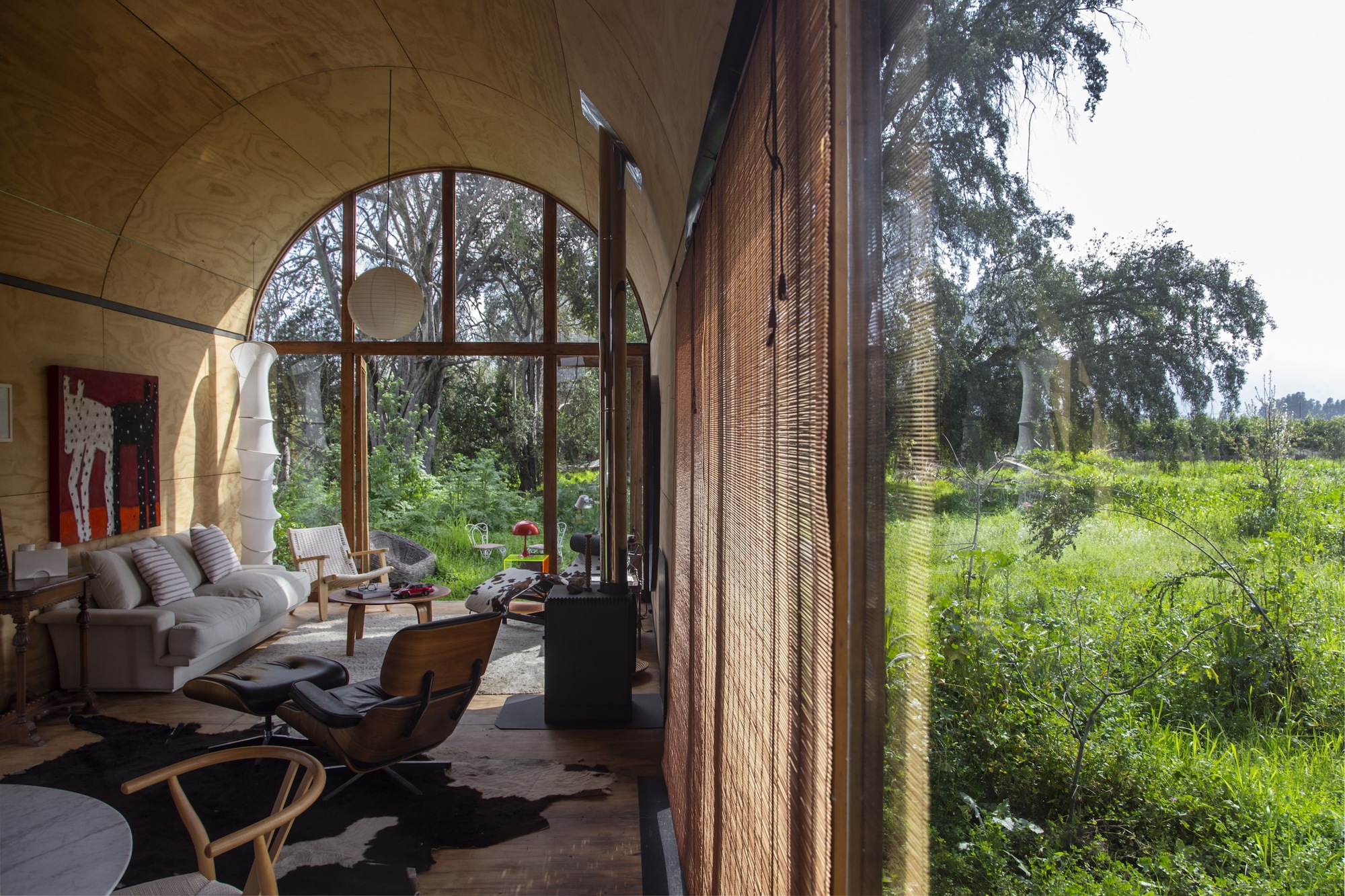 Living room seen past a woven bamboo blind, with plywood vault, arched window and a sunlit meadow outside