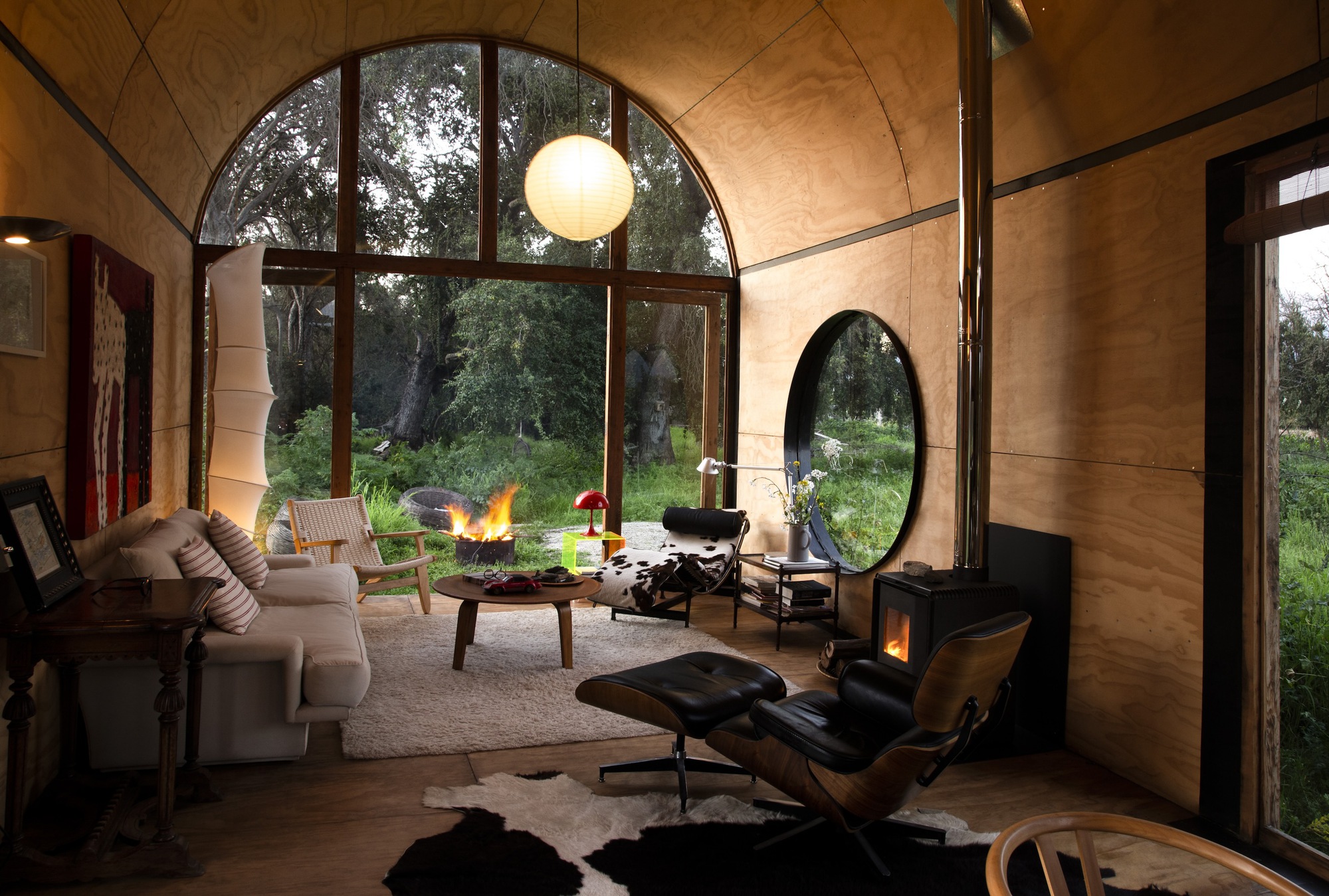 Living room at dusk with a lit wood stove, a fire burning in the garden and a round window framing trees