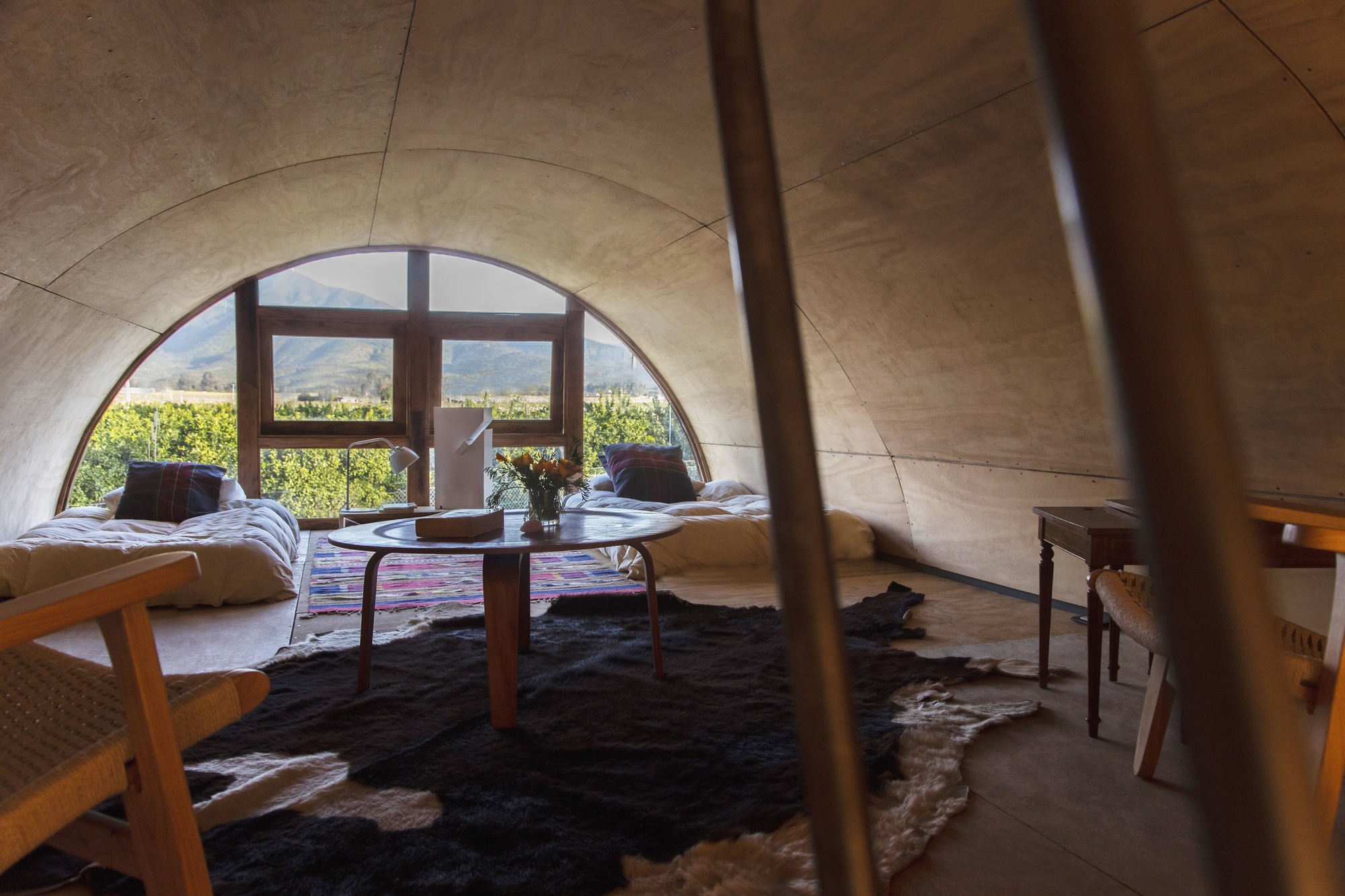 Plywood barrel-vaulted sleeping loft with floor beds, a round table and an arched window framing vineyards and mountains in daylight