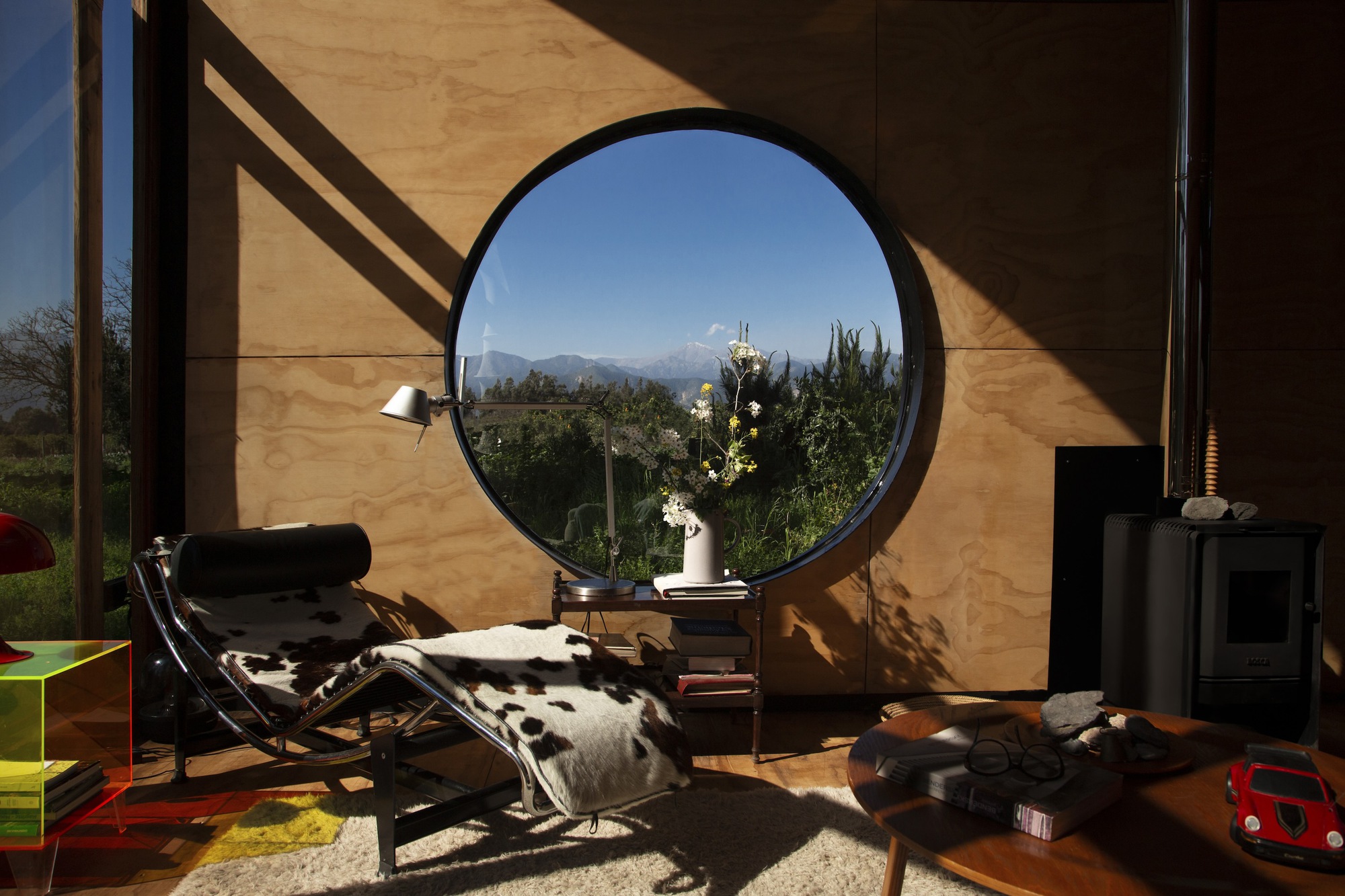Round window in a plywood wall framing mountains, with a leather and cowhide chaise in raking afternoon sun