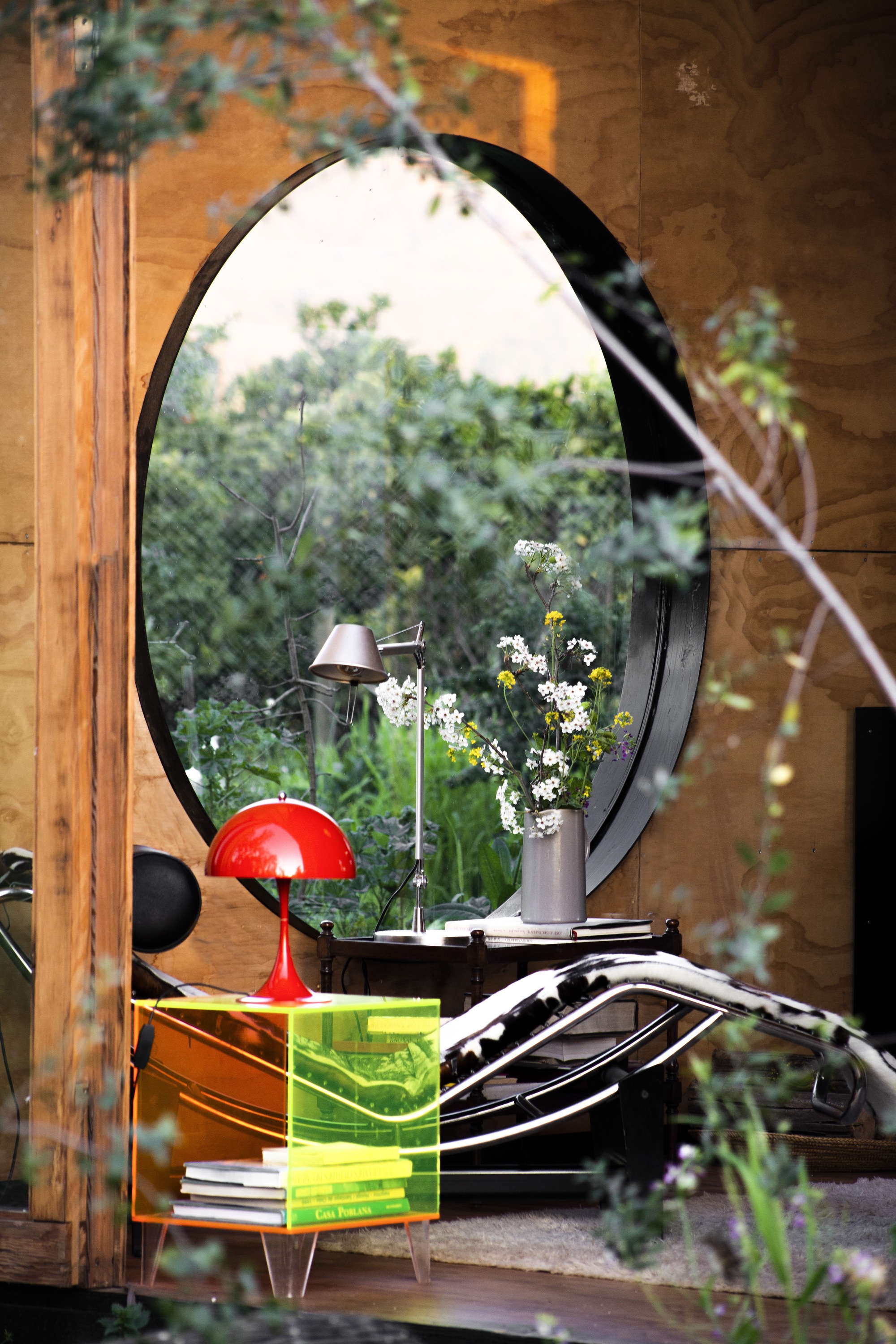 Oval window seen through leafy branches, with a red lamp on a translucent acrylic side table and a vase of white flowers