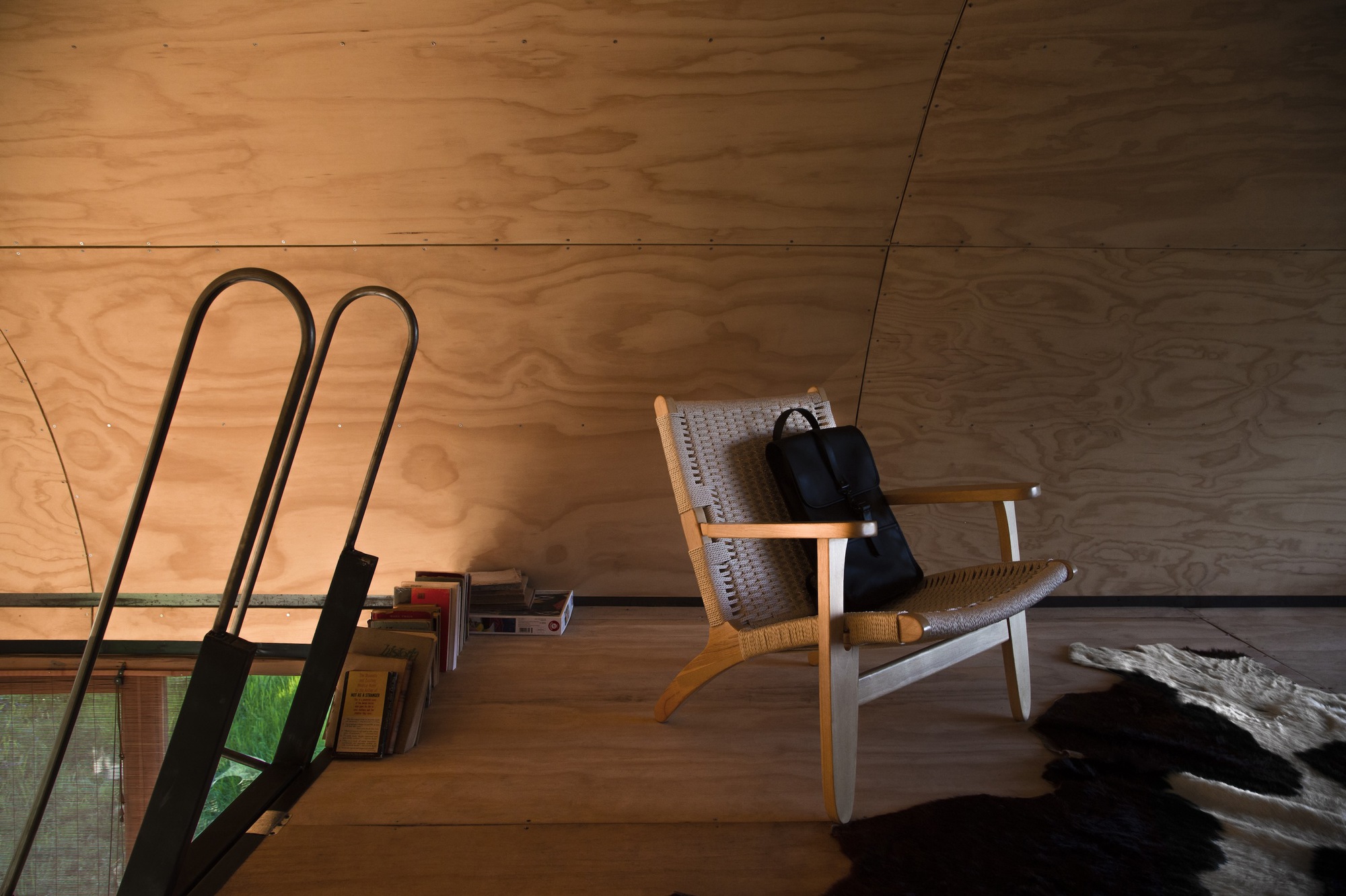 Woven-seat wooden armchair with a black bag beside a stack of books and a steel ladder under the curved plywood ceiling