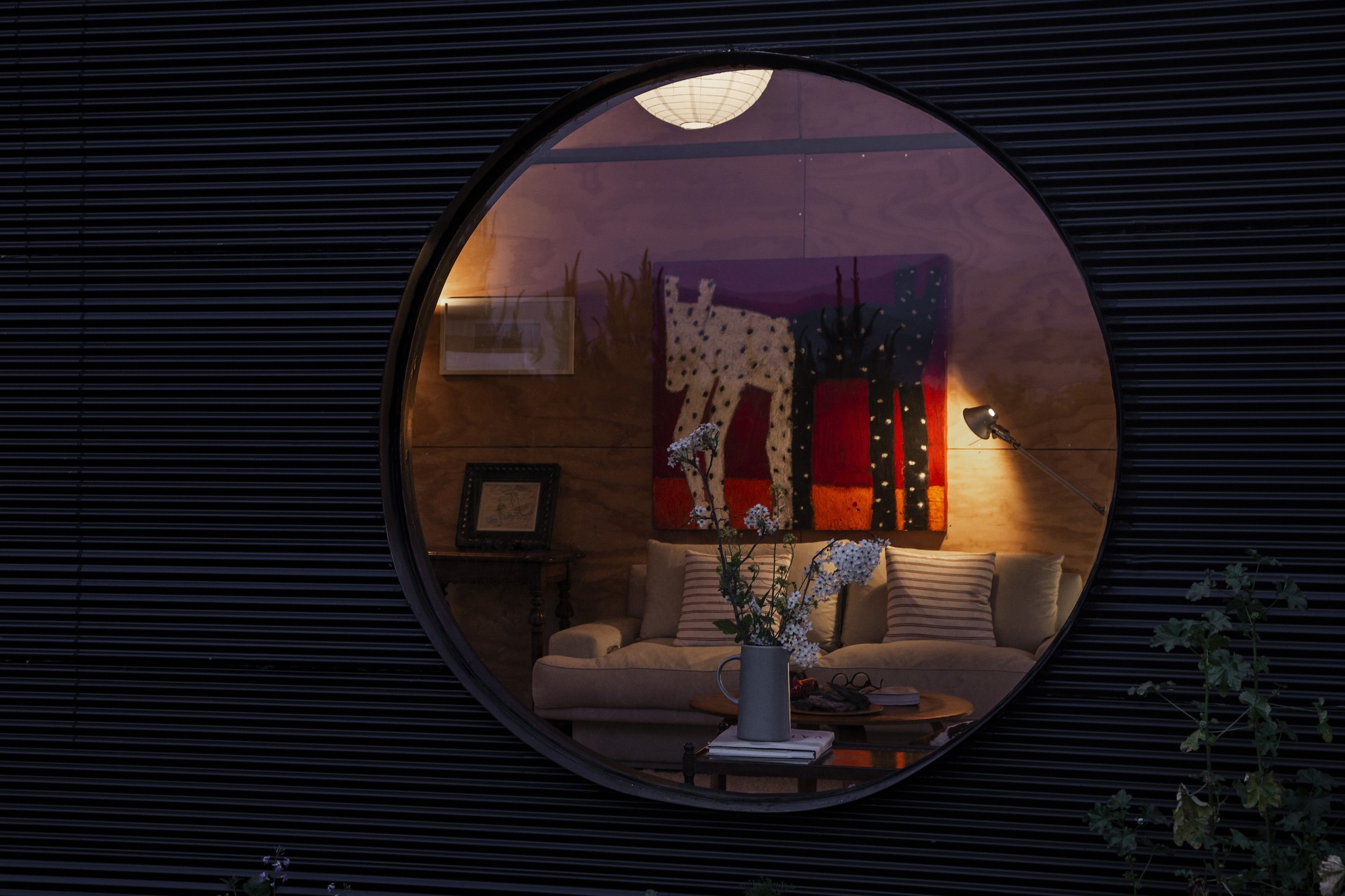 Round window cut into dark slatted cladding, framing a lit sofa, abstract painting and vase of white flowers at dusk