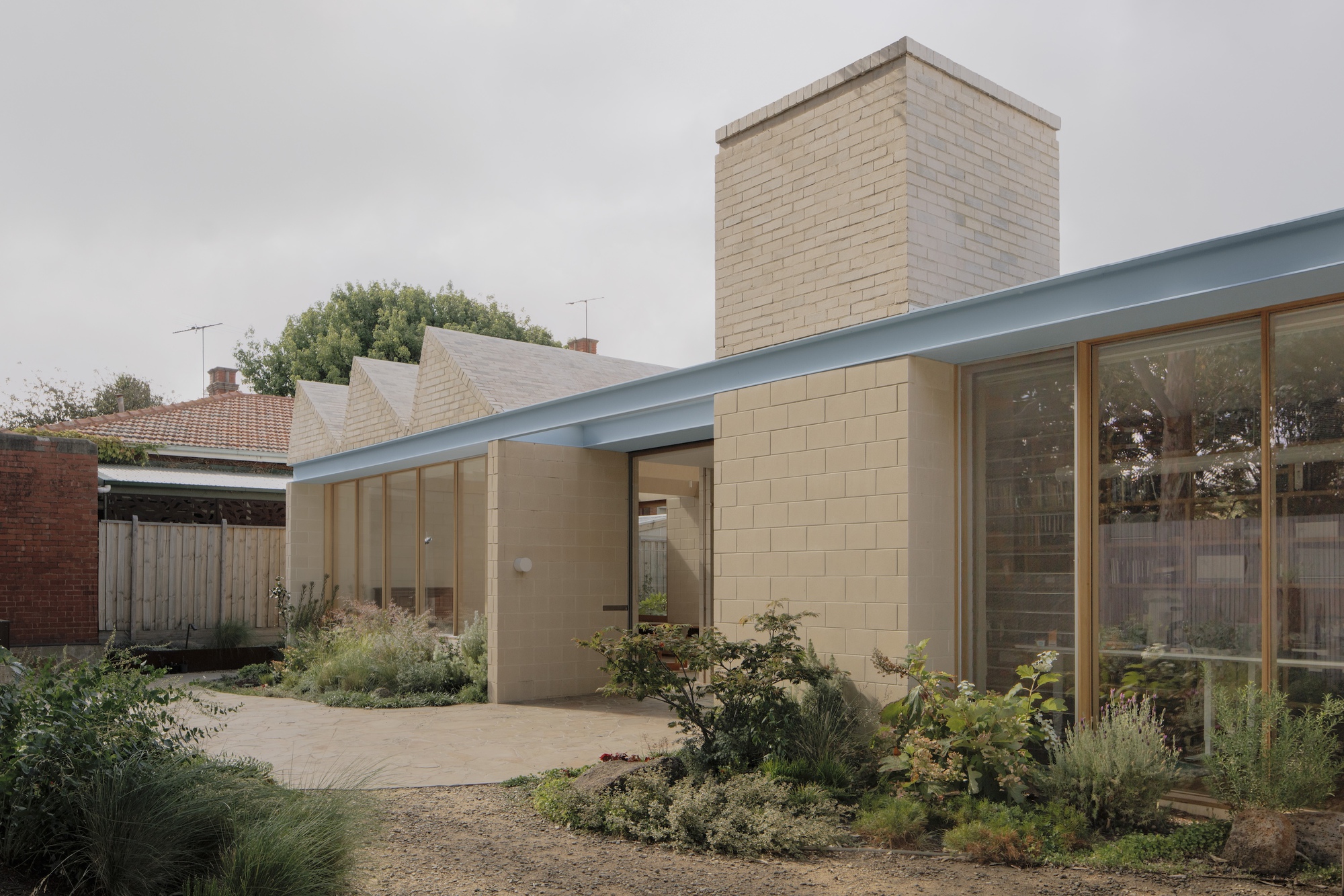 Garden-side view of the block chimney, pale blue roof edge and timber-framed glazing beside sawtooth roofs on a grey day