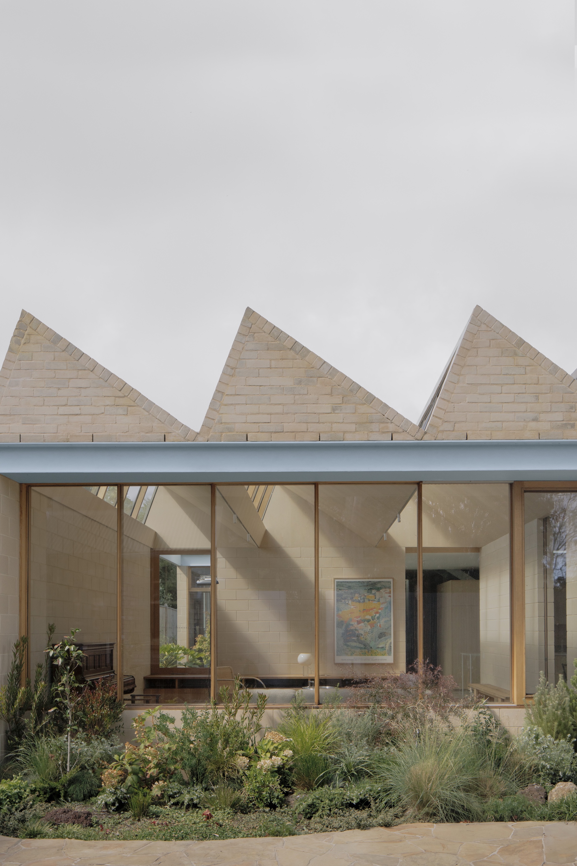 Three sawtooth gables of pale block above a glazed living space, with a planted garden bed in front under flat cloud