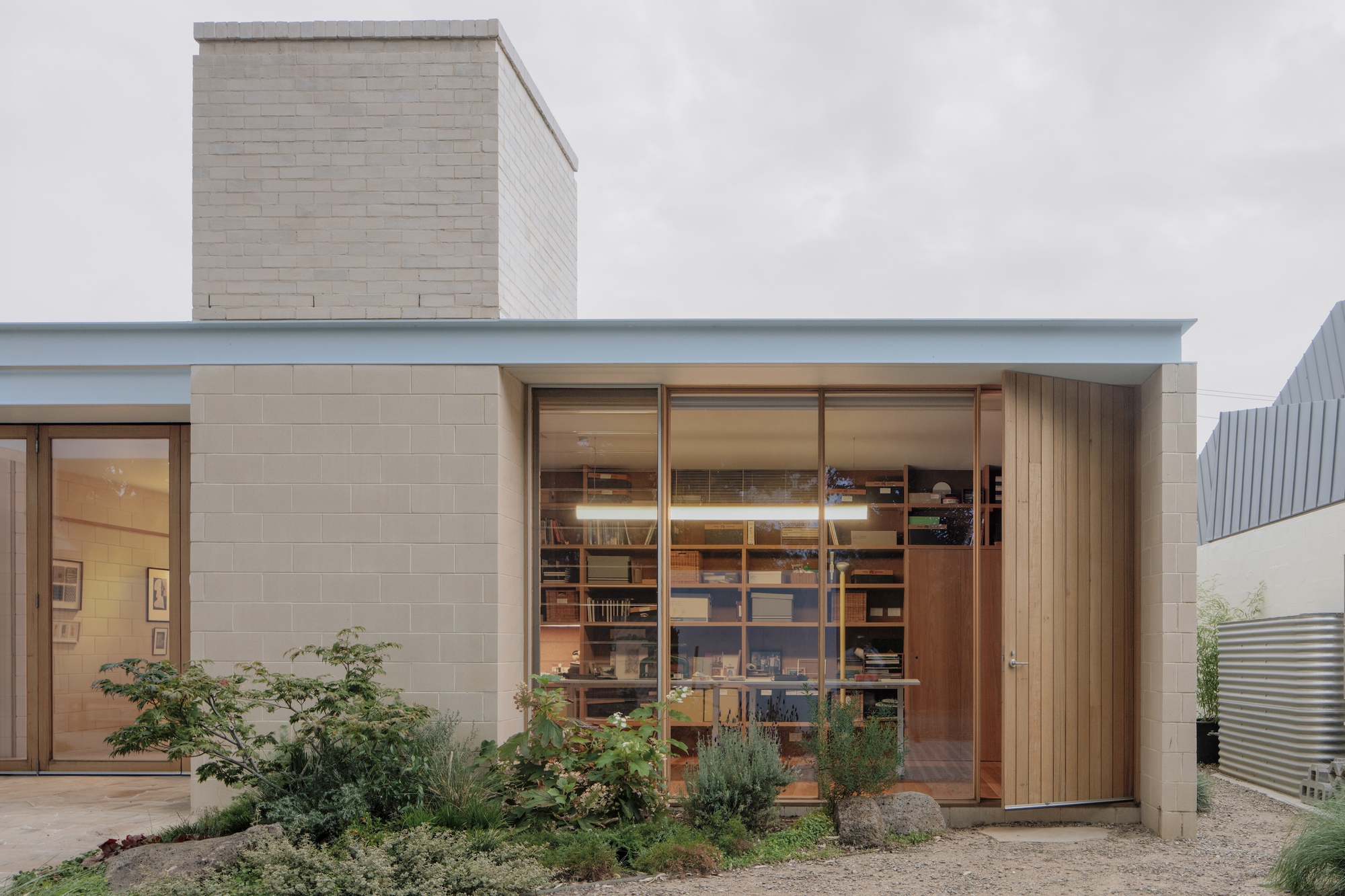 Study with shelves of books seen through full-height glazing, beside a vertical timber pivot door and a block chimney in dull light