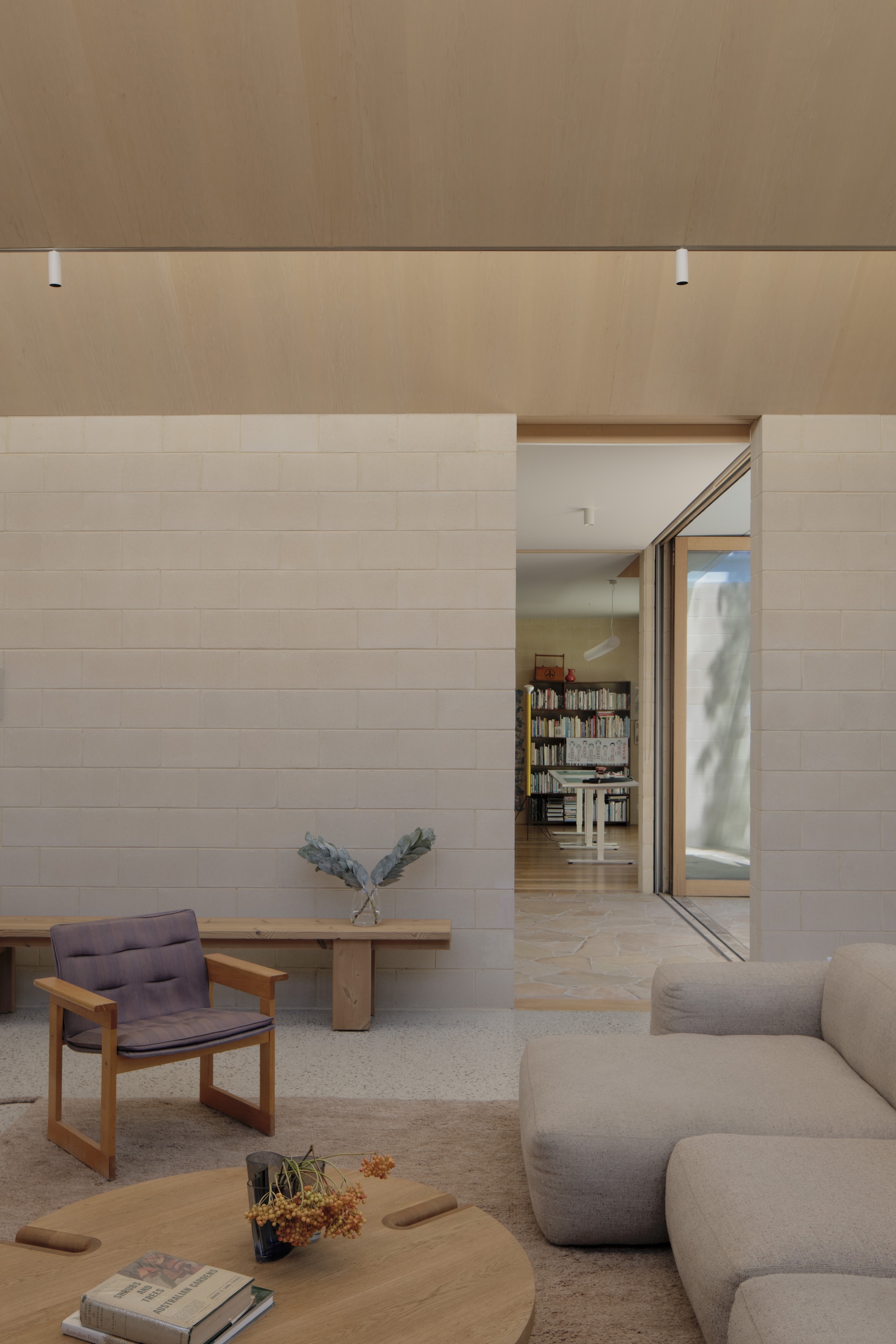 Living room with block wall, timber bench, wooden armchair and low sofa, looking through a doorway to a bookshelf beyond