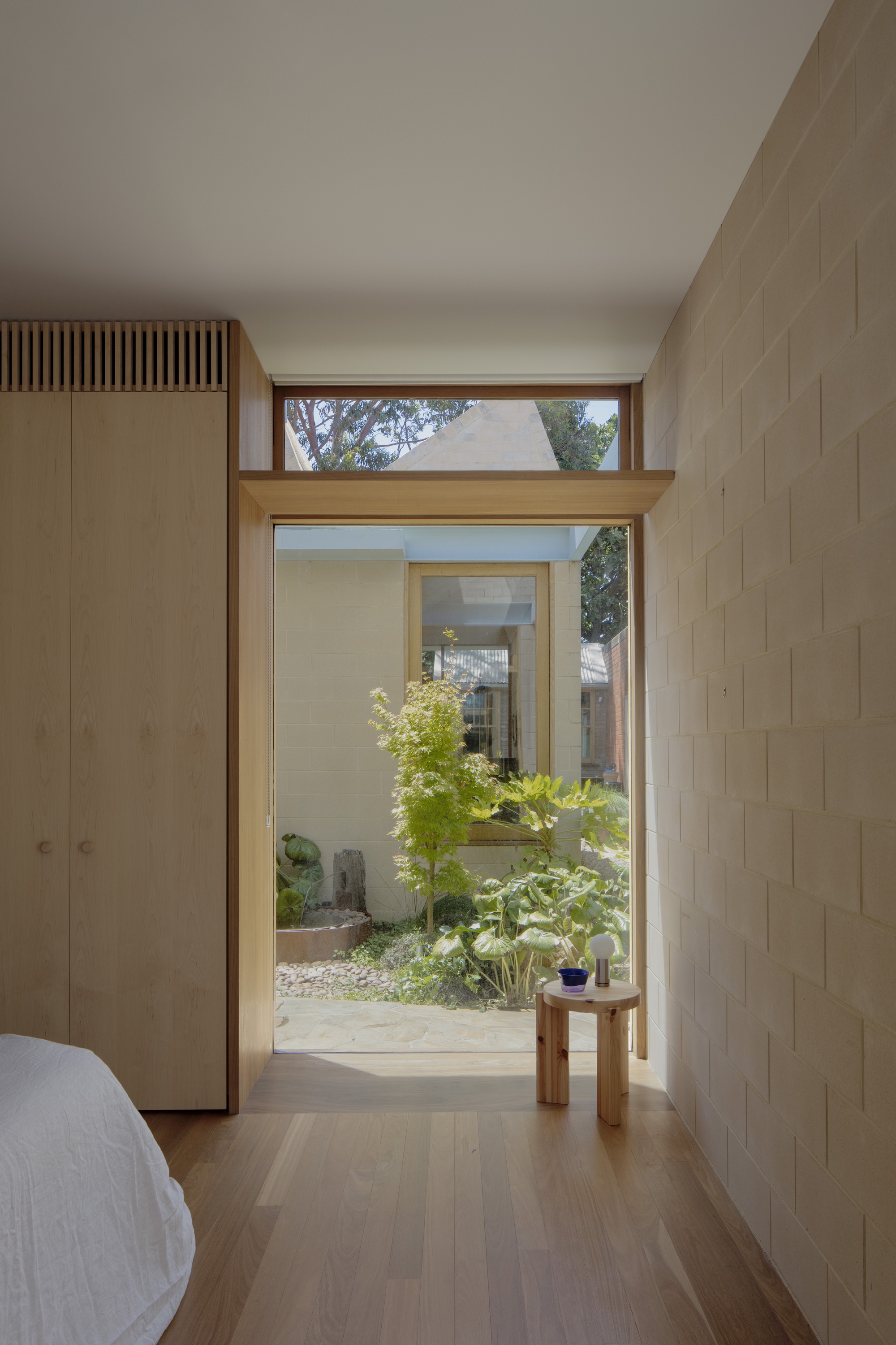 Bedroom doorway framing a courtyard with a maple and broad-leaved plants, beside timber wardrobe and block wall in afternoon sun