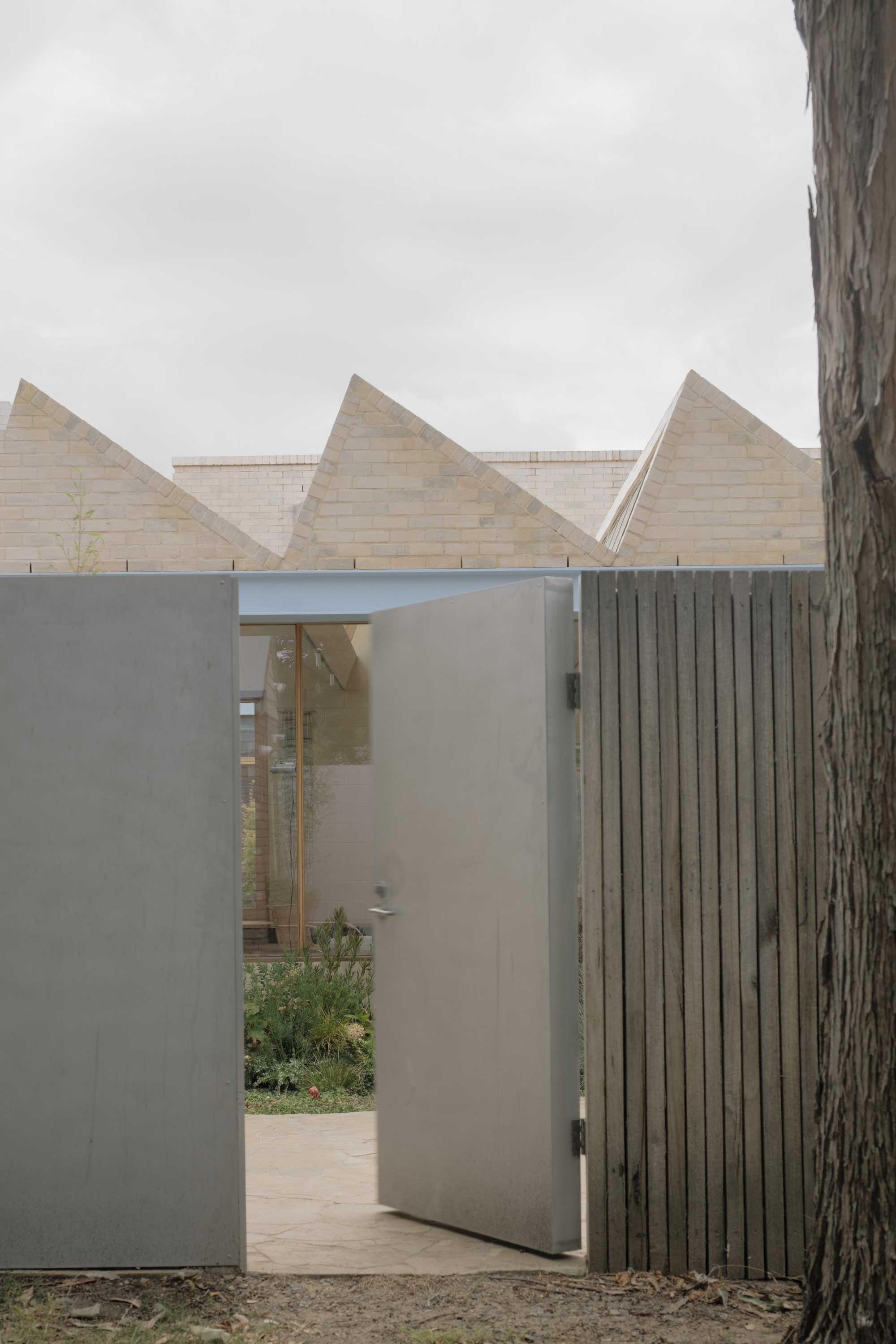 Open steel-clad gate door beside a timber fence, revealing a planted courtyard below pale brick sawtooth gables