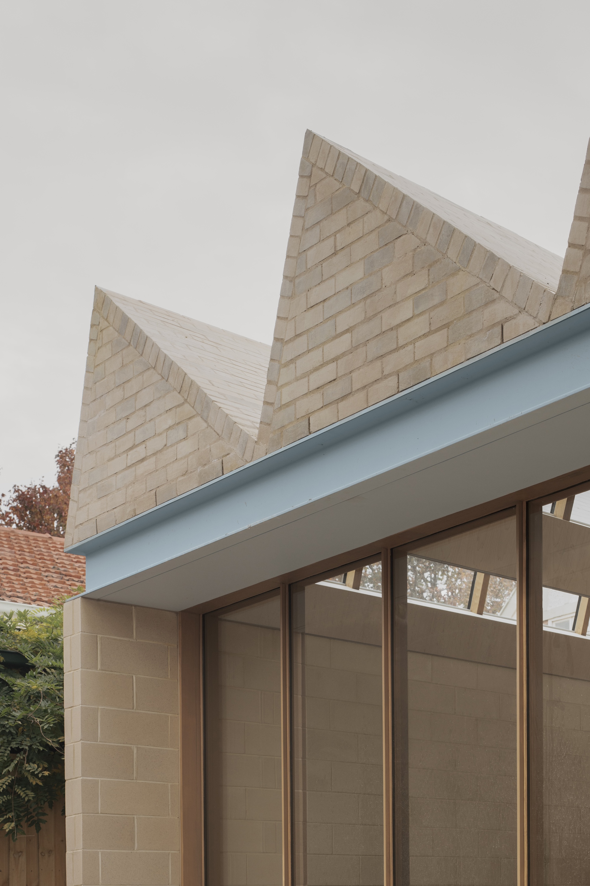 Close-up of pale brick sawtooth gables above a light blue steel fascia and timber-framed glazing, with autumn foliage behind