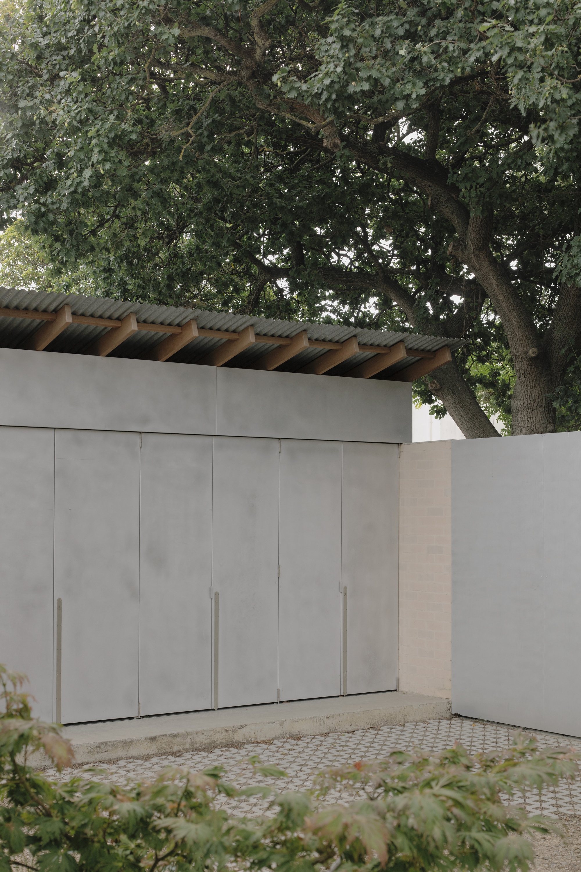 Grey panel doors with slim steel pulls under a corrugated roof with exposed timber rafters, shaded by a large oak