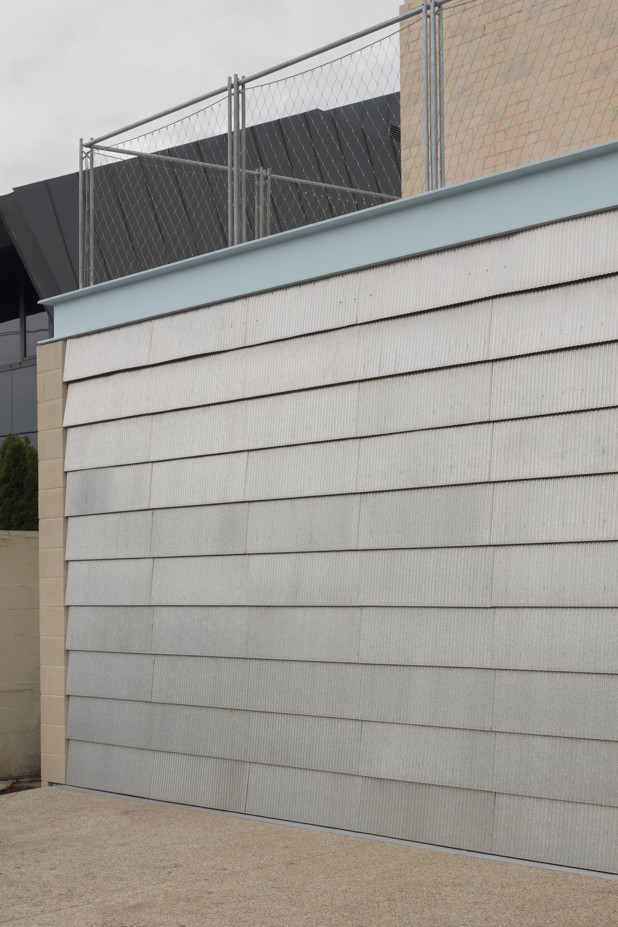 Overlapping ribbed fibre-cement cladding boards below a pale blue fascia, with chain-link fencing and a dark roof behind under overcast sky