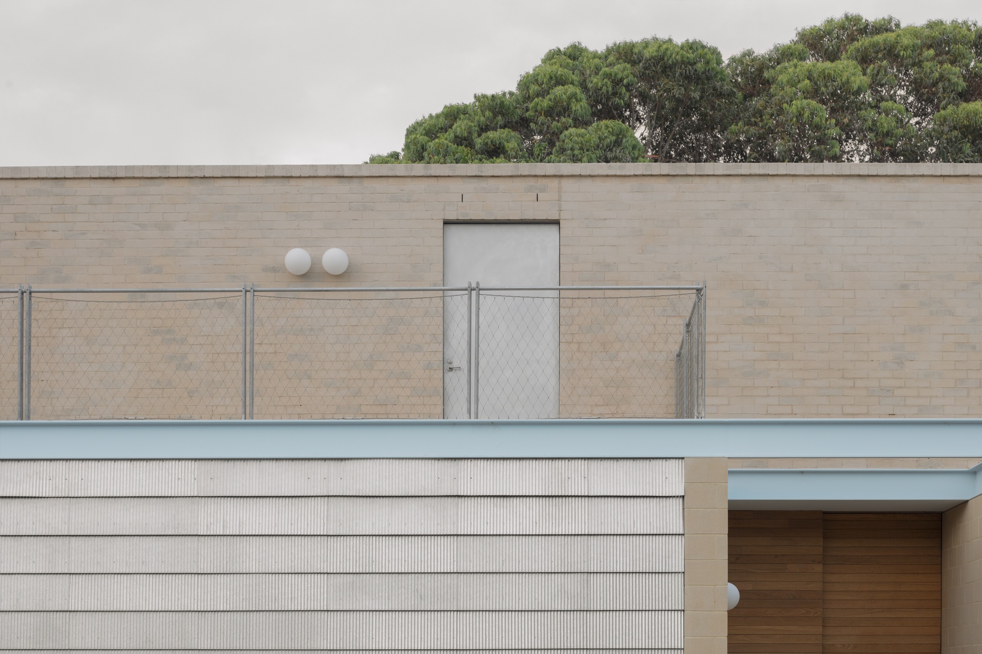 Rooftop brick wall with a grey door, chain-link fence and two globe lights, with a tree canopy rising behind in grey light