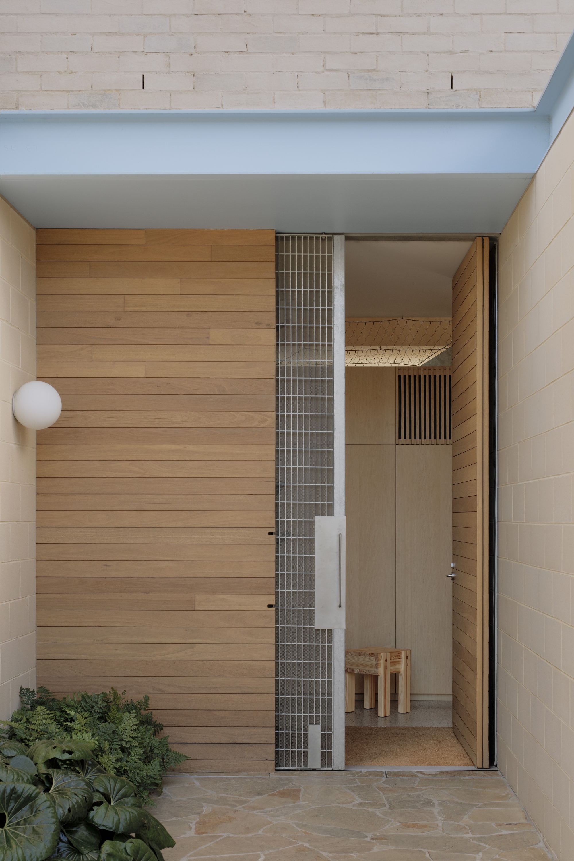 Entry with timber-clad pivot door, galvanised mesh panel and globe light, set beside a bed of broad-leaved plants on flagstone paving