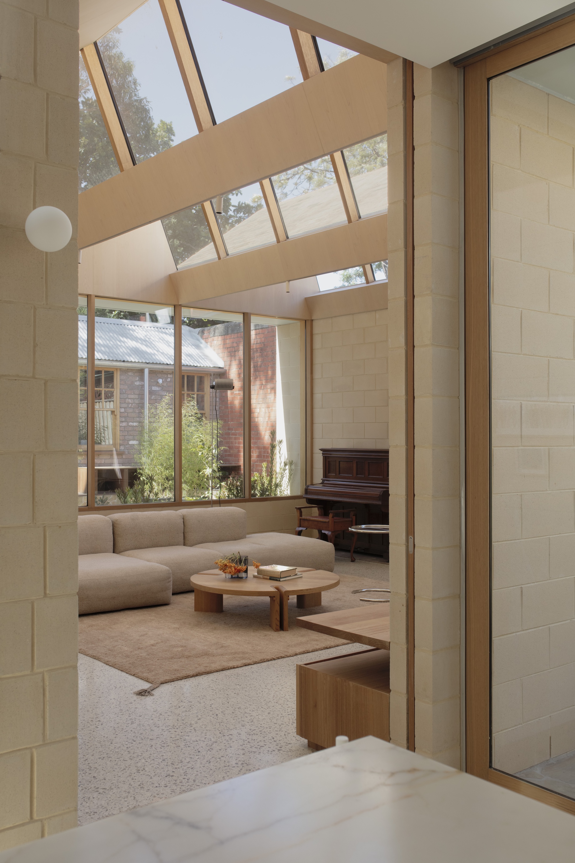 Living room seen past a block wall, with a glazed skylight, low sofa and upright piano, and neighbouring brick houses through the glass