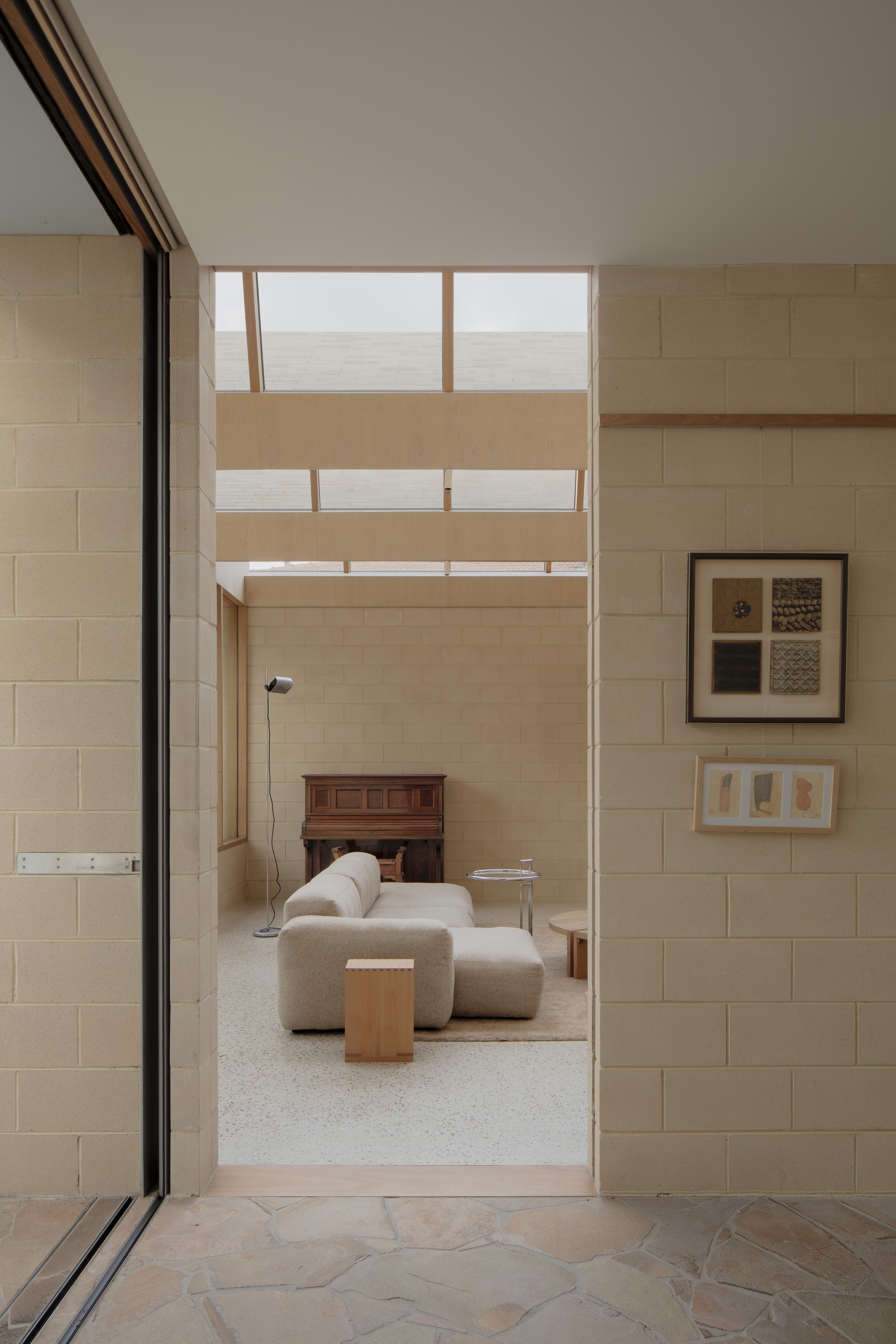 Doorway view through cream block walls to a sofa, piano and skylight, with framed artworks on the wall and flagstone underfoot