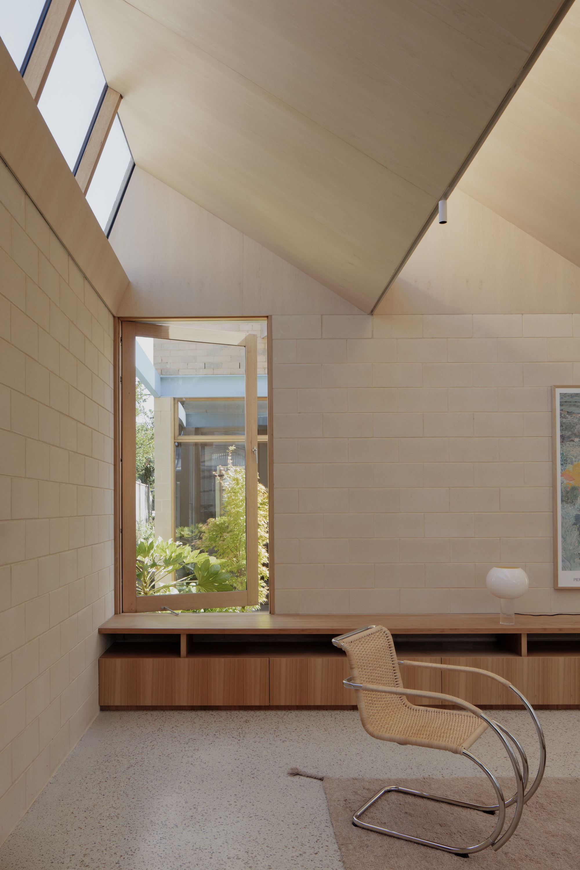 Corner of a room with a pitched plywood ceiling, pivot window onto greenery, timber joinery bench and a cantilevered cane chair in daylight