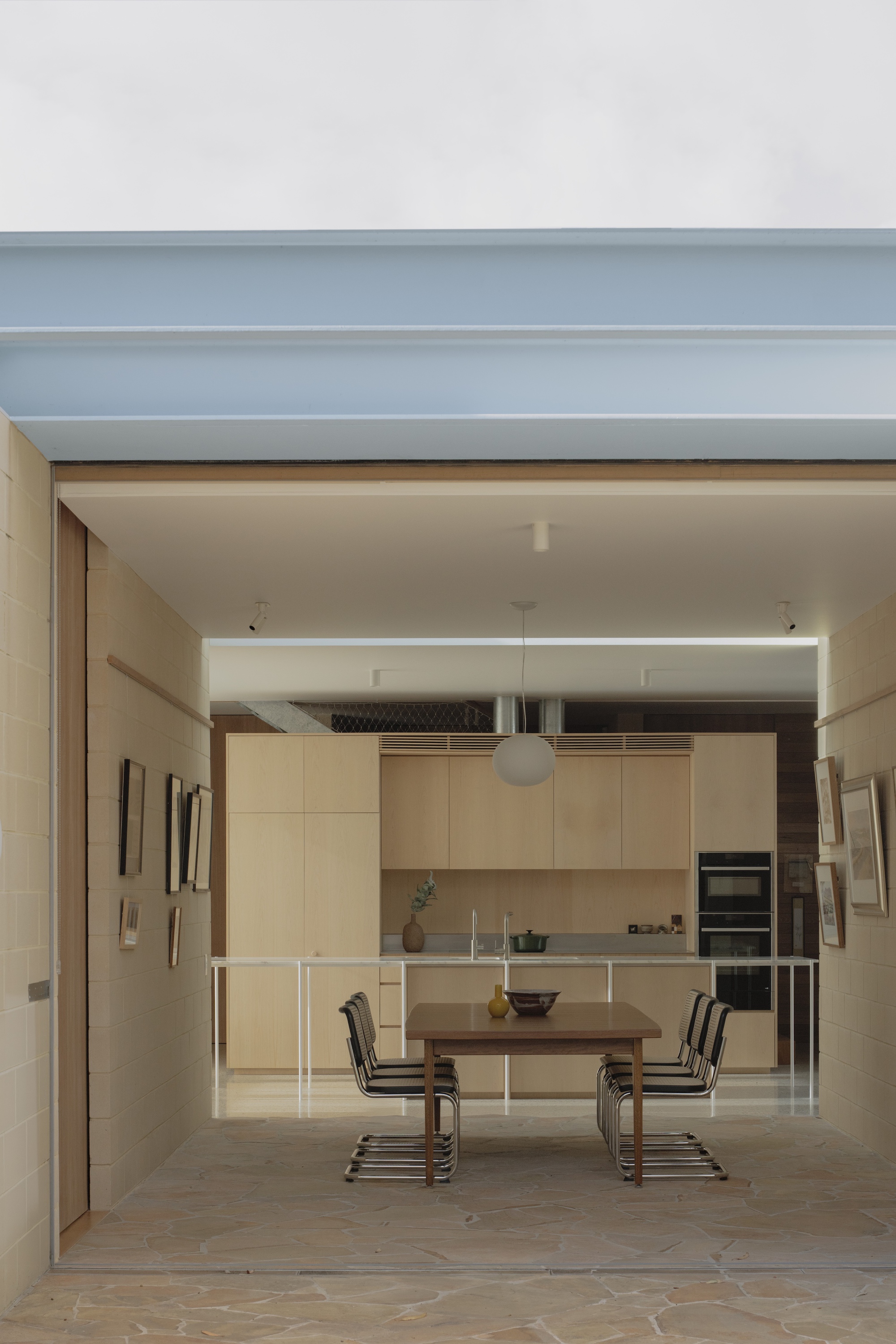 Open dining area framed by a pale blue steel beam, with a timber table, wire-frame chairs and oak kitchen beyond