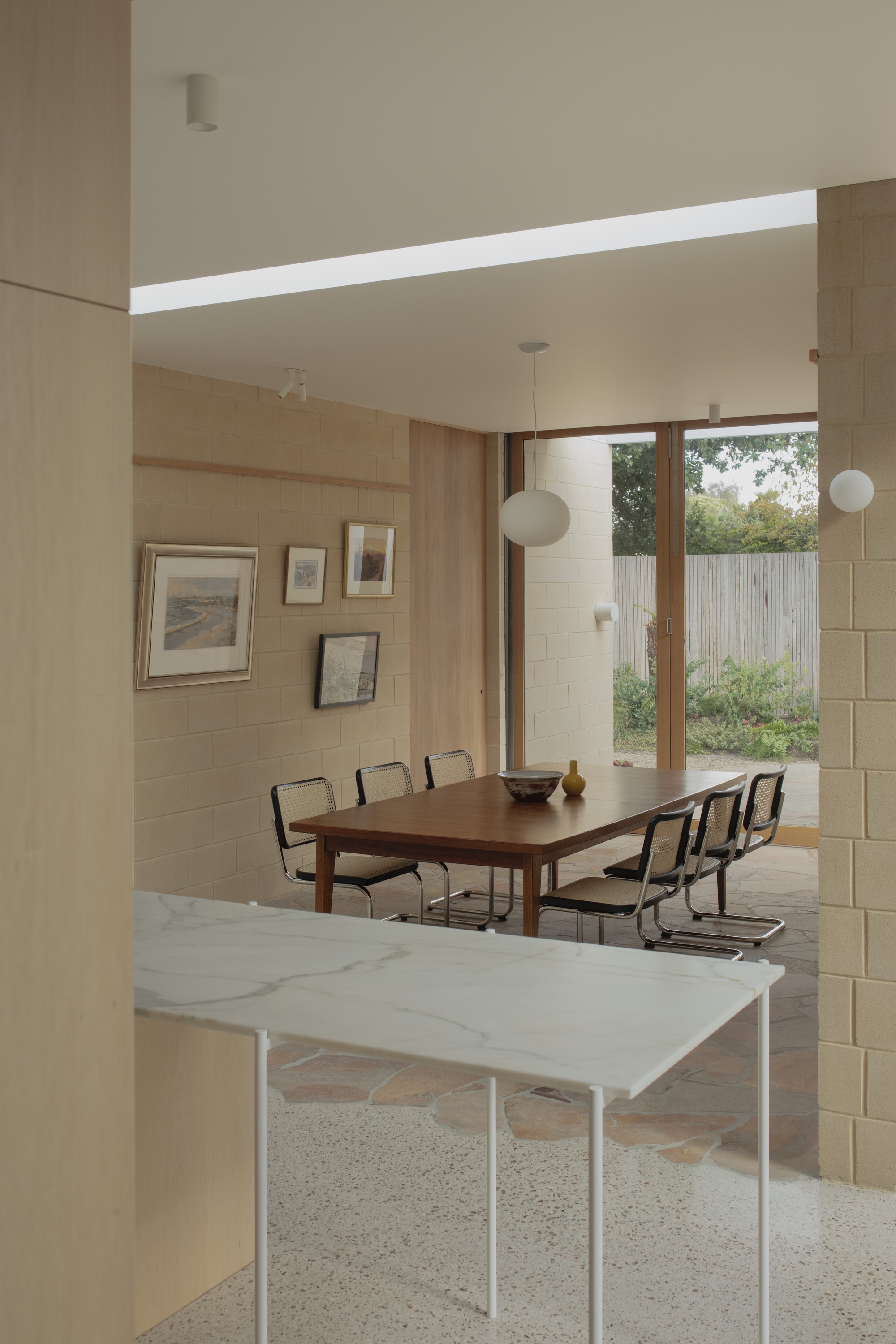 Marble-topped bench on slim white legs in the foreground, with a long dining table and garden glimpsed through timber-framed glass