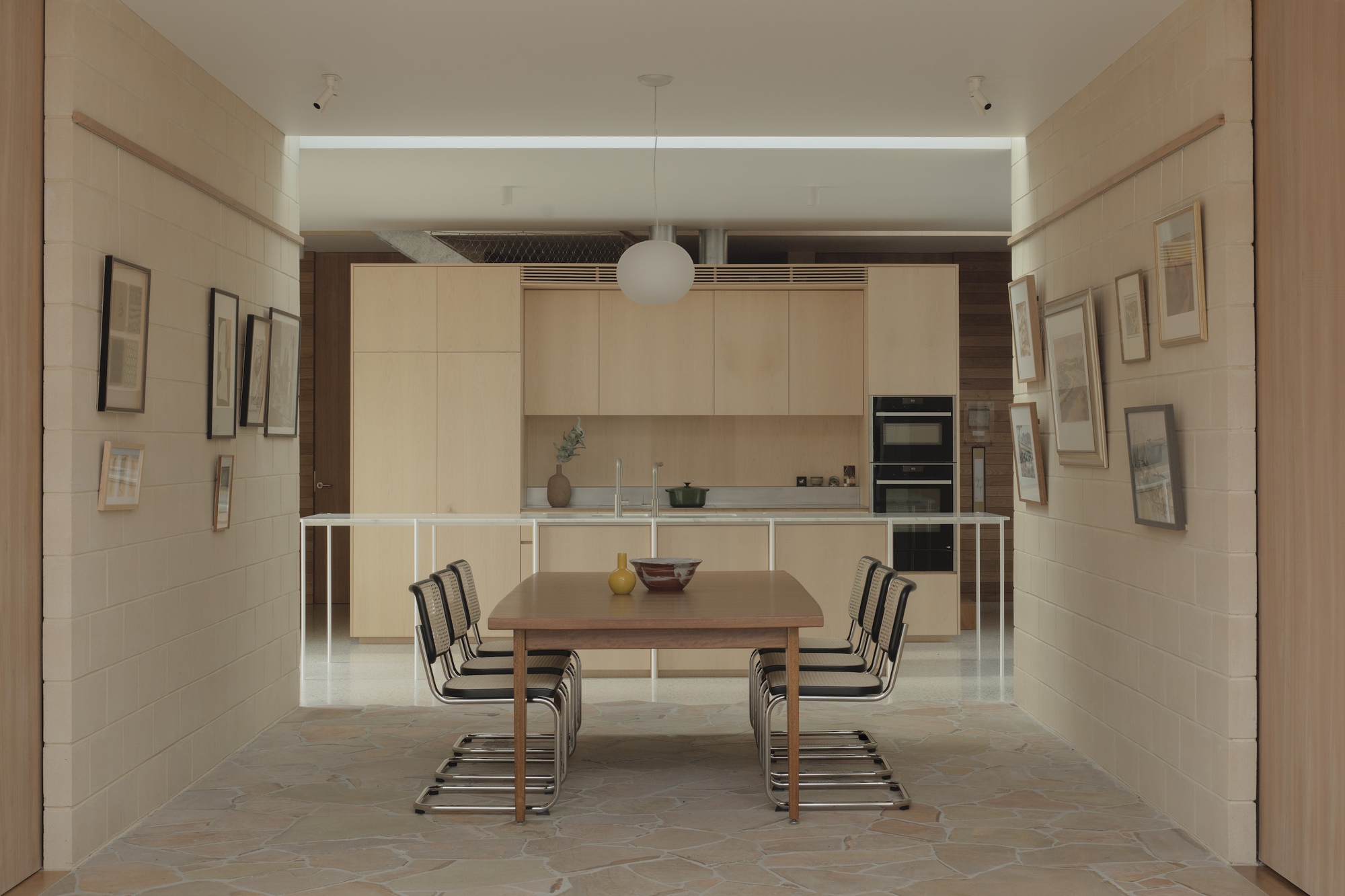 Symmetrical view down a corridor of framed artworks on block walls to a dining table and oak kitchen wall