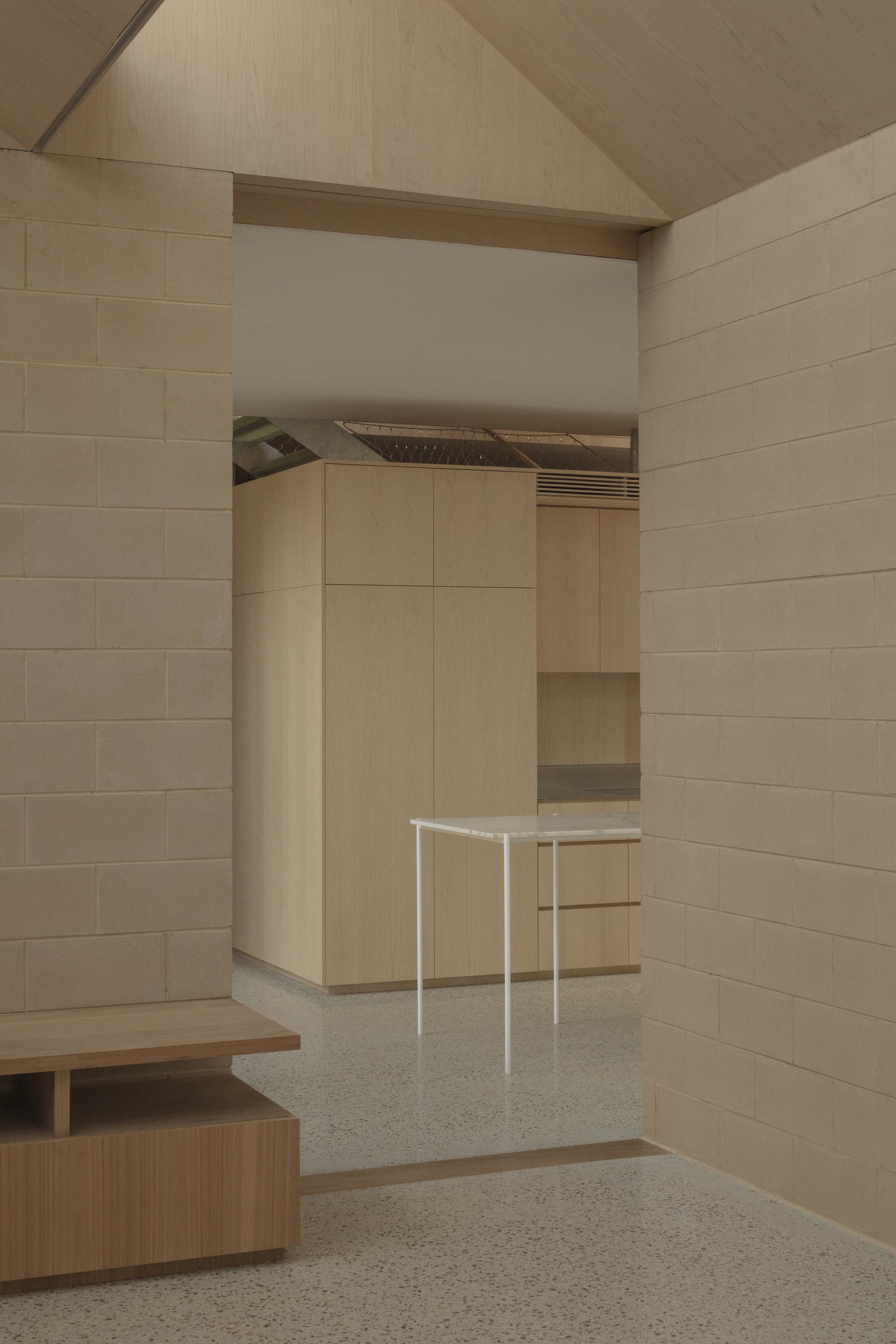 Pale concrete block walls and a pitched timber ceiling framing a doorway toward a white-legged bench and oak cabinetry
