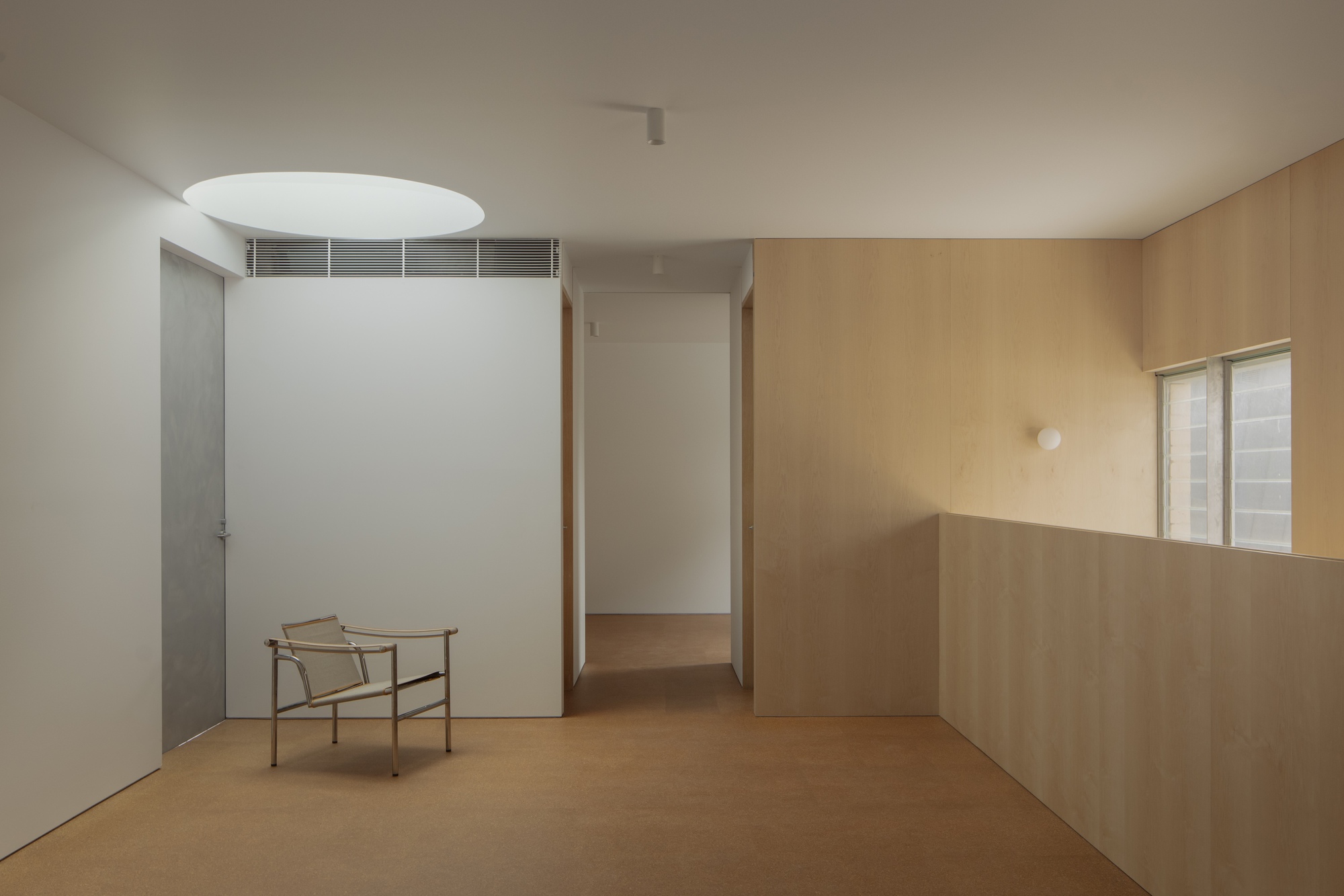 Upper room with a round skylight, a lone steel-framed chair on a cork floor and birch plywood walls in dim daylight