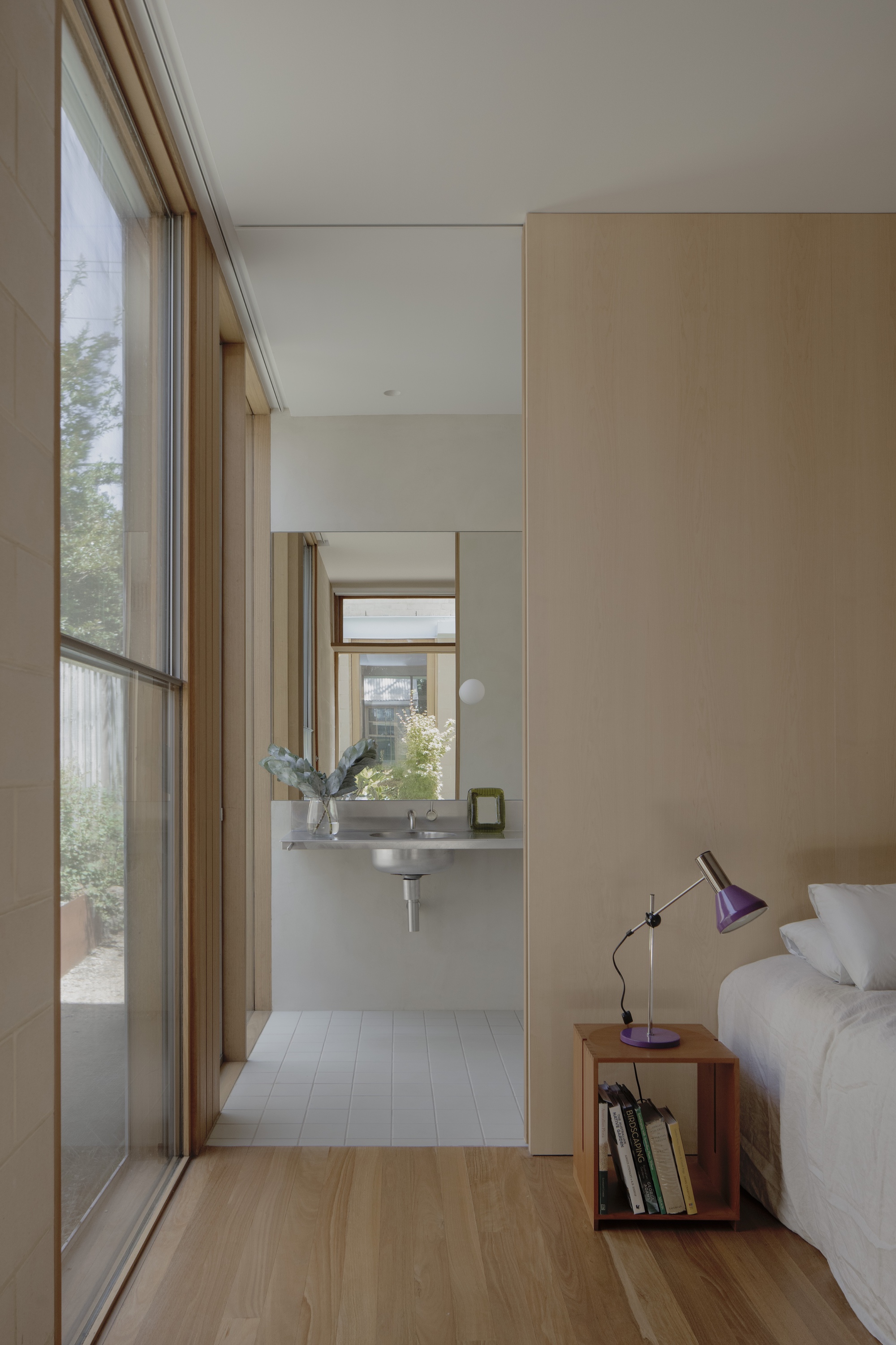 Bedroom corner with a floor-to-ceiling glass wall, a purple desk lamp on a timber crate and a mirrored basin beyond