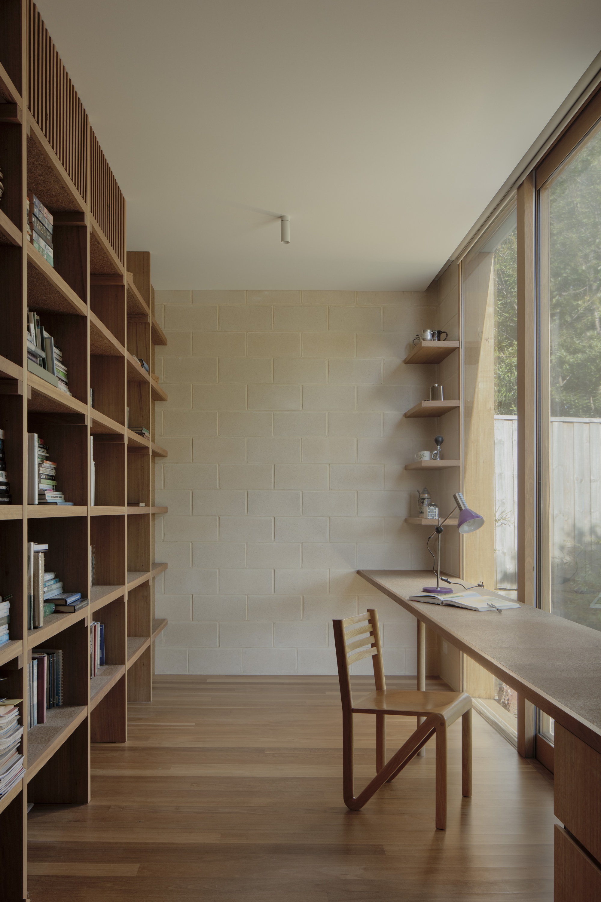 Study with a full-height timber bookshelf, a long desk and wooden chair facing a concrete block wall and tall garden window
