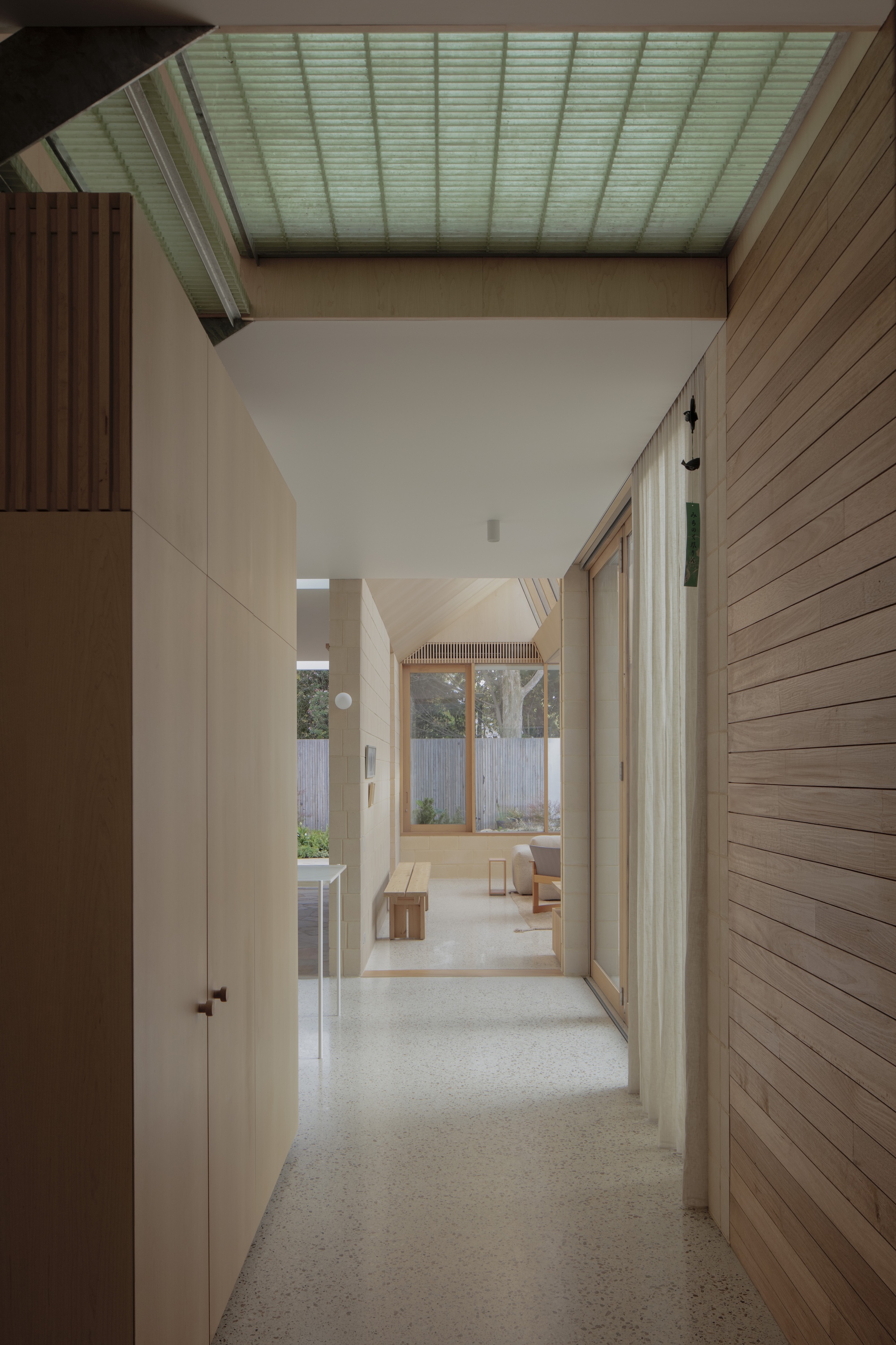 Corridor with a green-tinted skylight, timber cladding and plywood cabinets leading toward a garden window and a bench in soft light