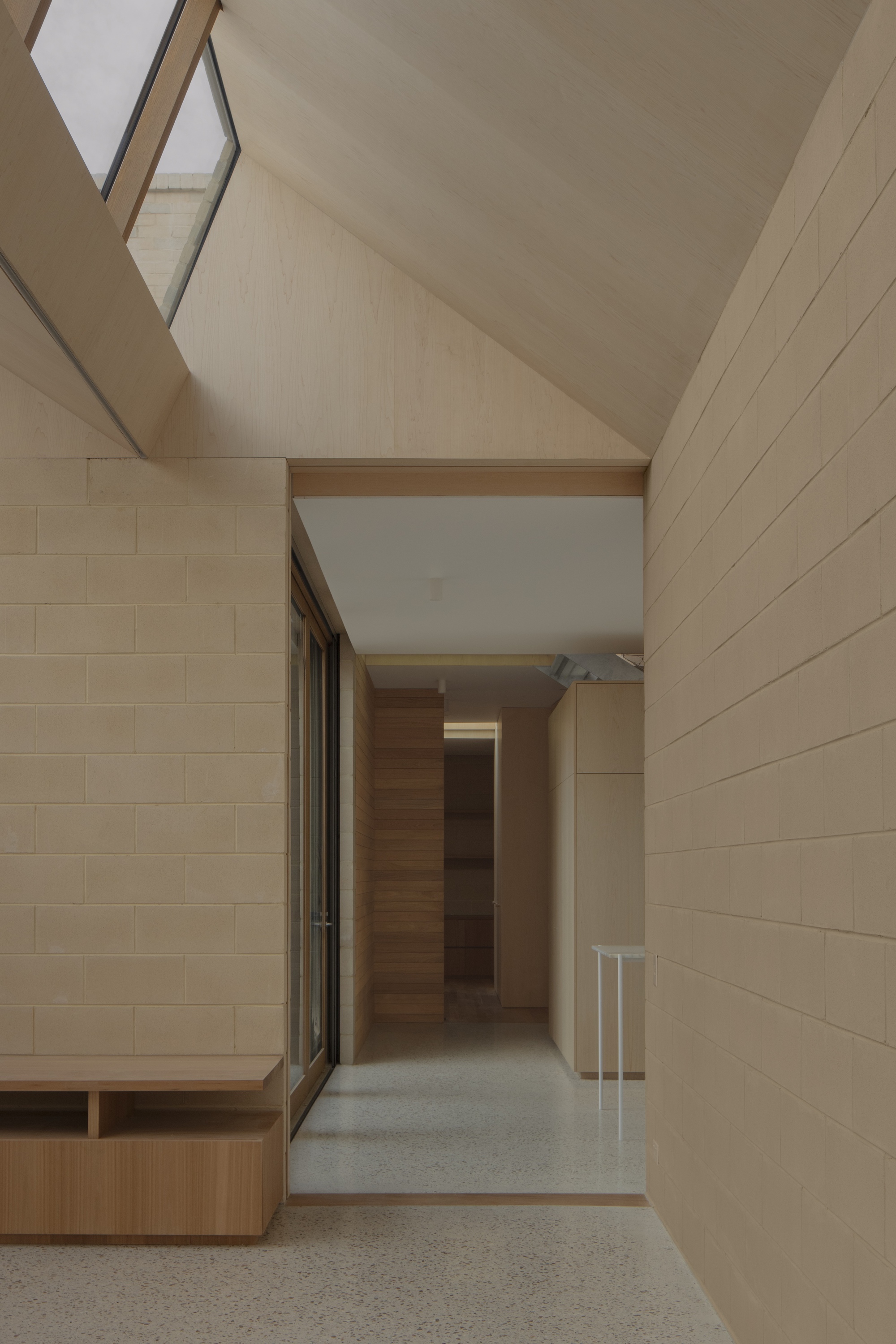 Hallway of pale concrete block walls and a pitched plywood ceiling with a skylight, over a speckled terrazzo floor