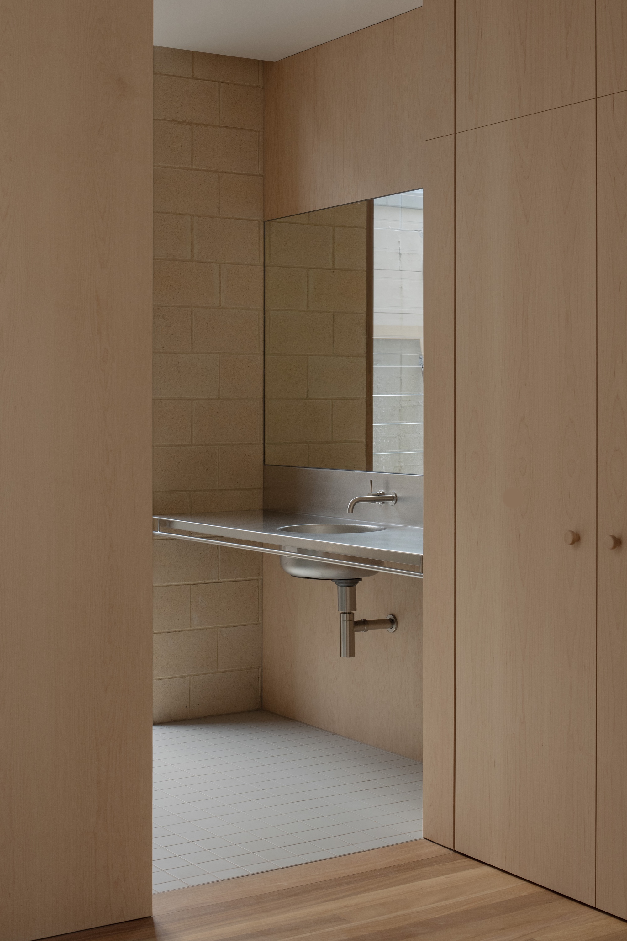 Stainless steel basin and wall tap seen through a doorway between oak panelled walls, with concrete block behind and a mirror above