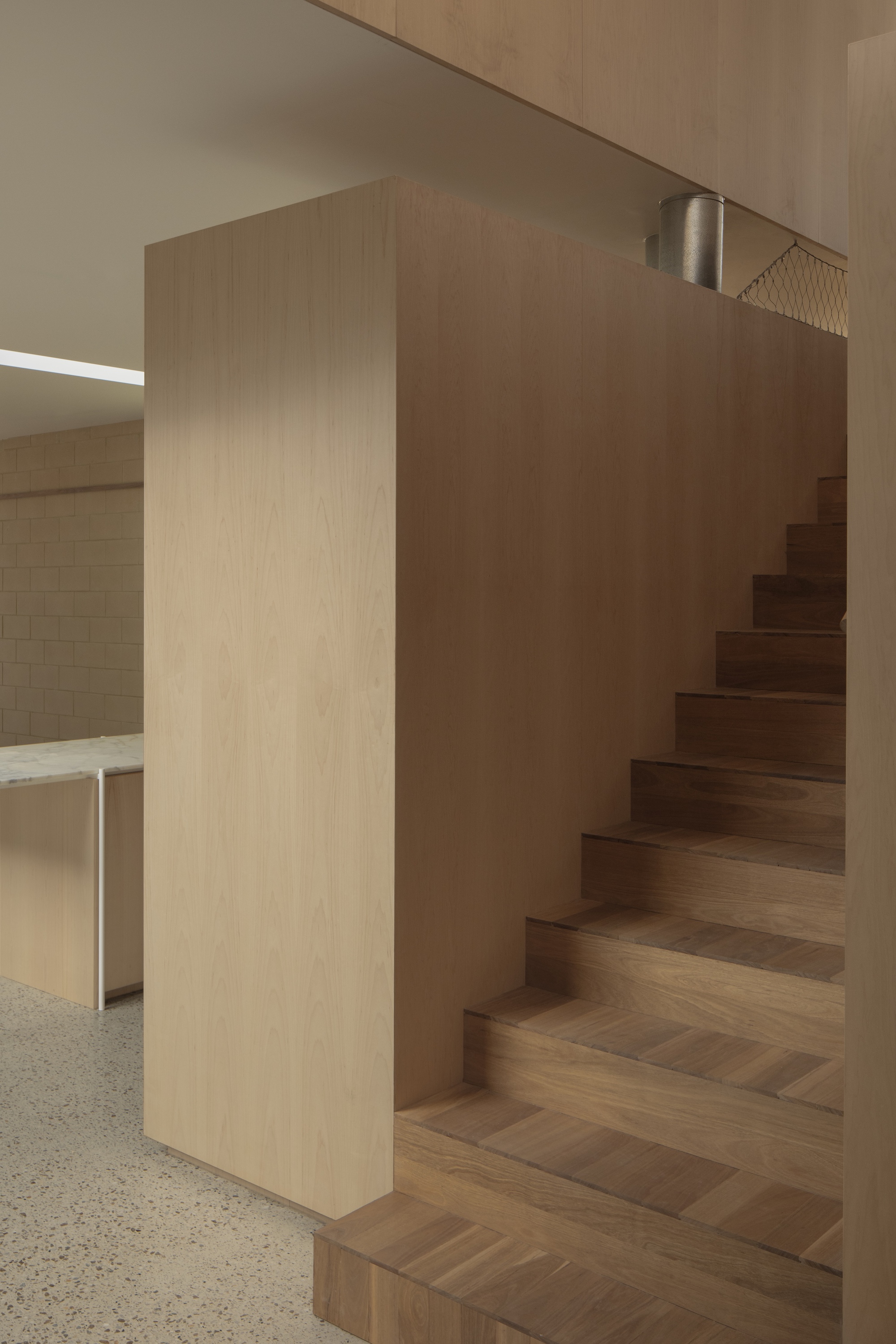 Oak clad stair volume with timber treads beside a terrazzo floor and a kitchen counter lit by a ceiling strip